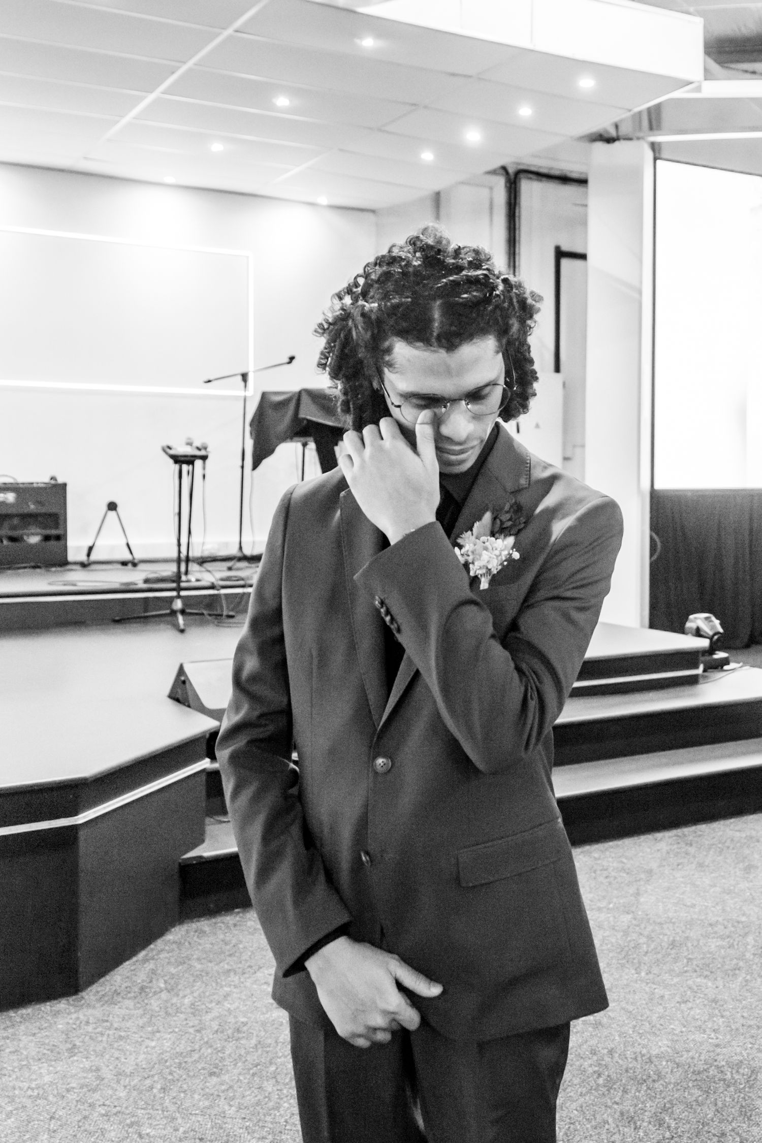 A person in a dark suit and boutonniere stands in what appears to be a church or event space, looking down while smiling.