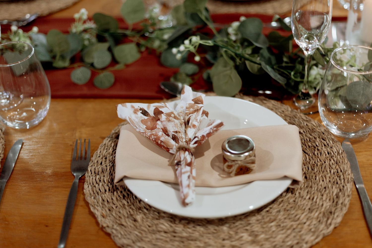 Rustic table setting with woven charger, white plate, floral napkin and eucalyptus garland on wooden surface.
