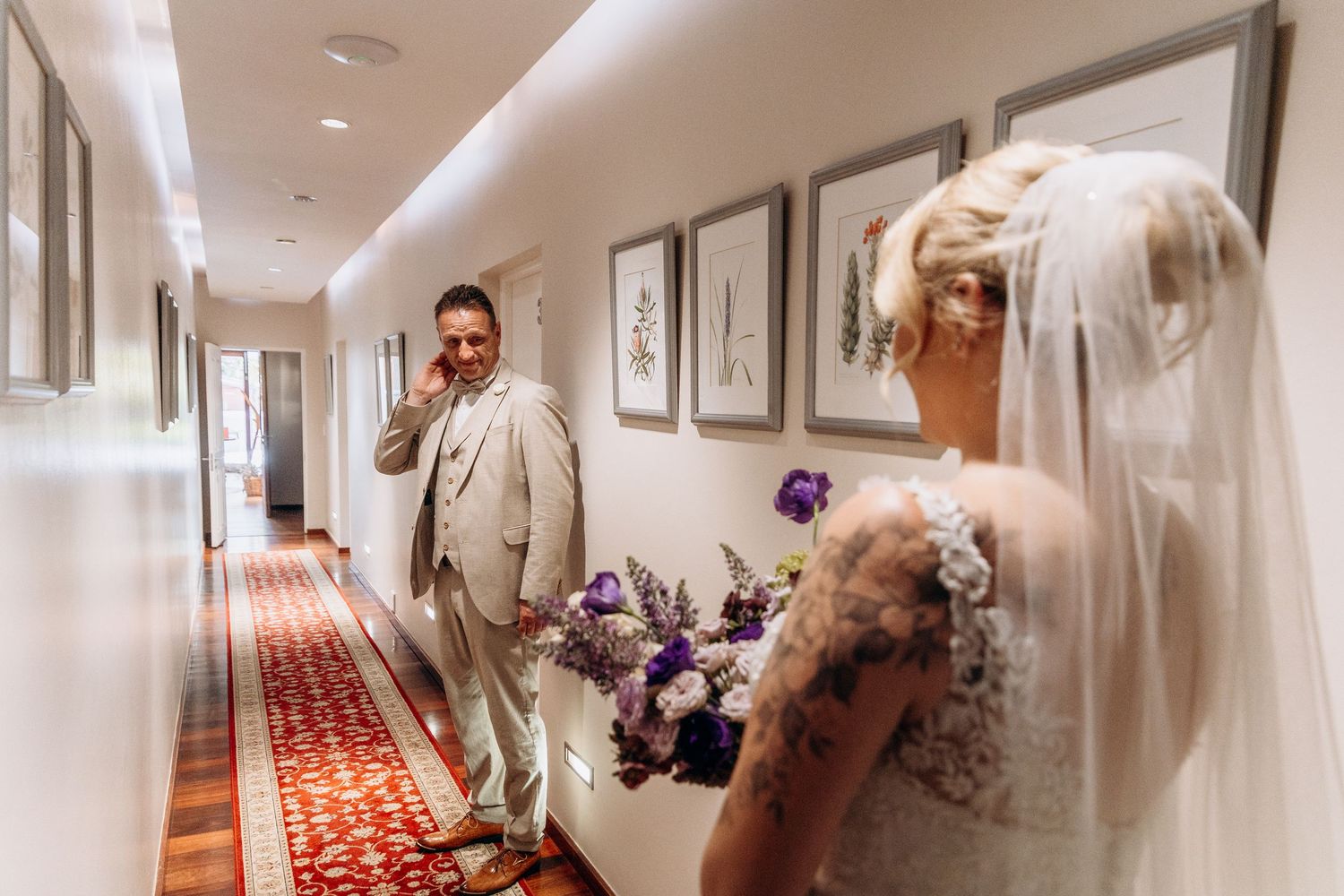 Emotional groom in beige suit reacts to seeing tattooed bride in white gown holding purple bouquet in hallway.