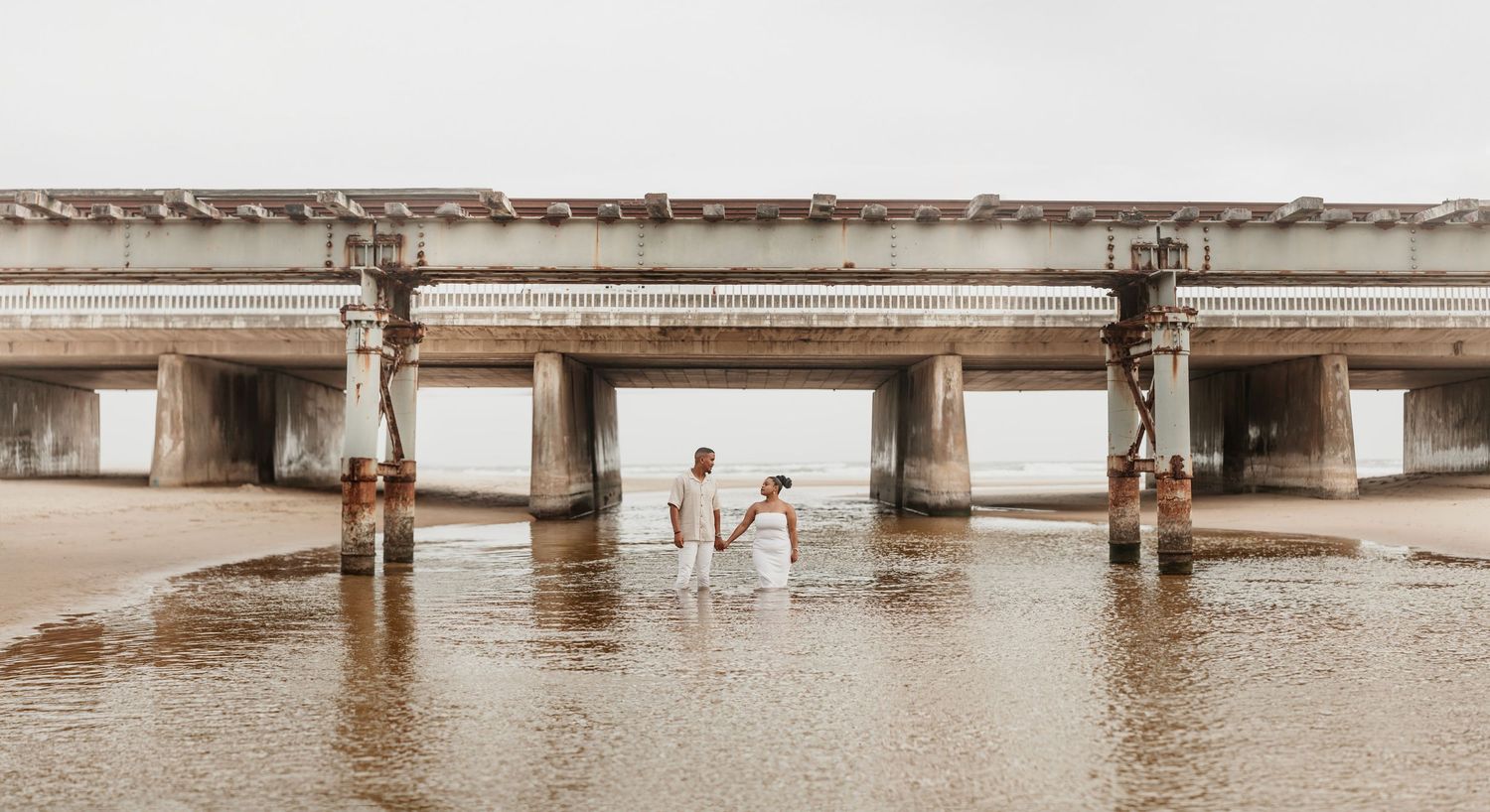 A couple holds hands while walking in shallow water beneath a concrete pier structure on a misty day.