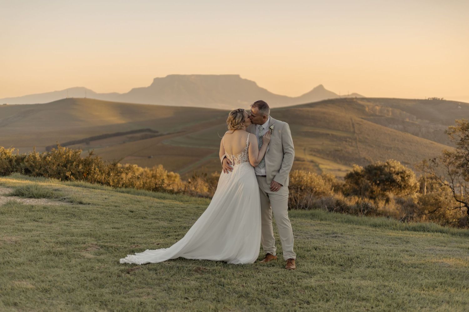 Bride and groom share a romantic kiss at sunset with rolling hills and Table Mountain in the background.