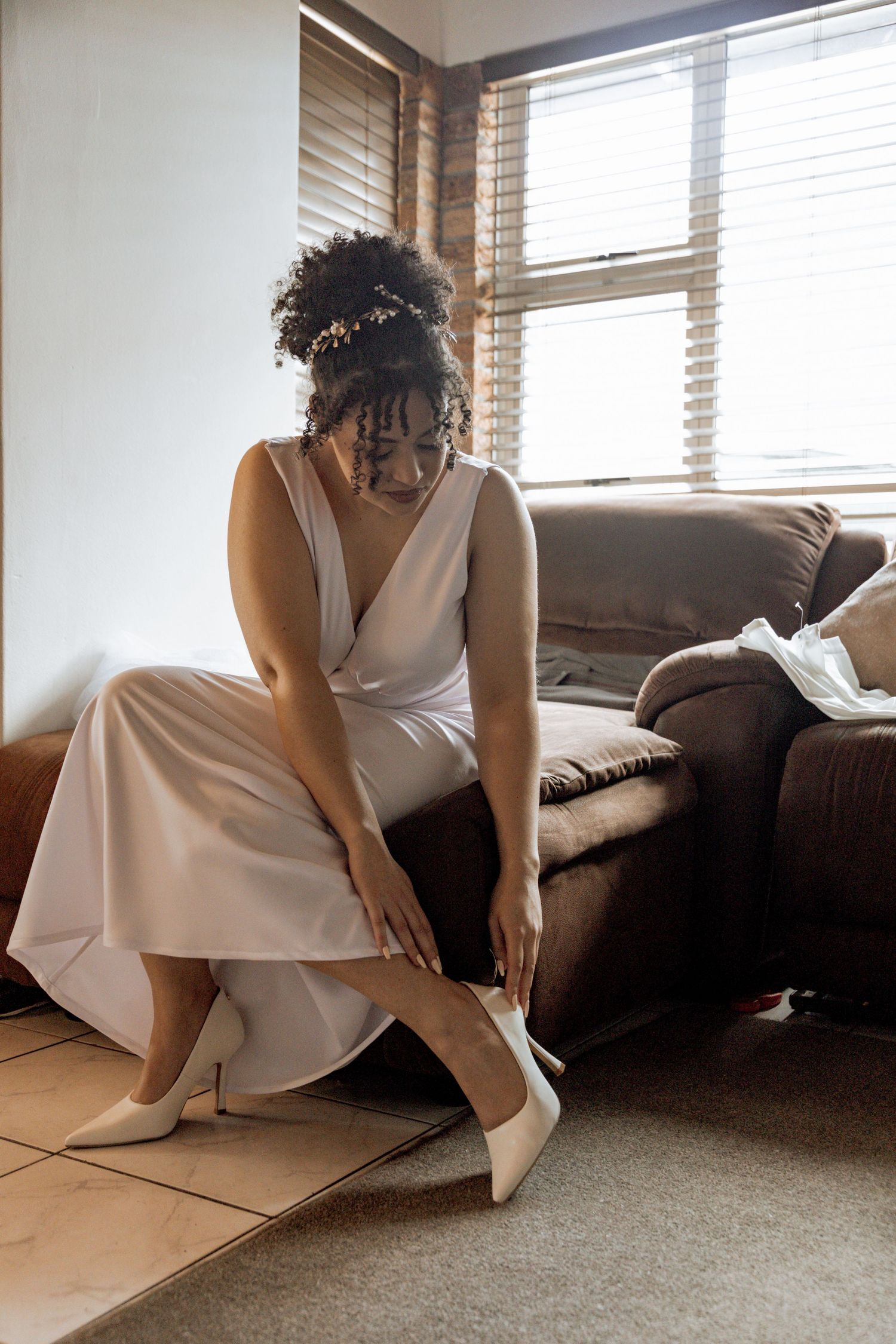 A person wearing an elegant white dress and heels sits on a brown couch in a sunlit room with window blinds.