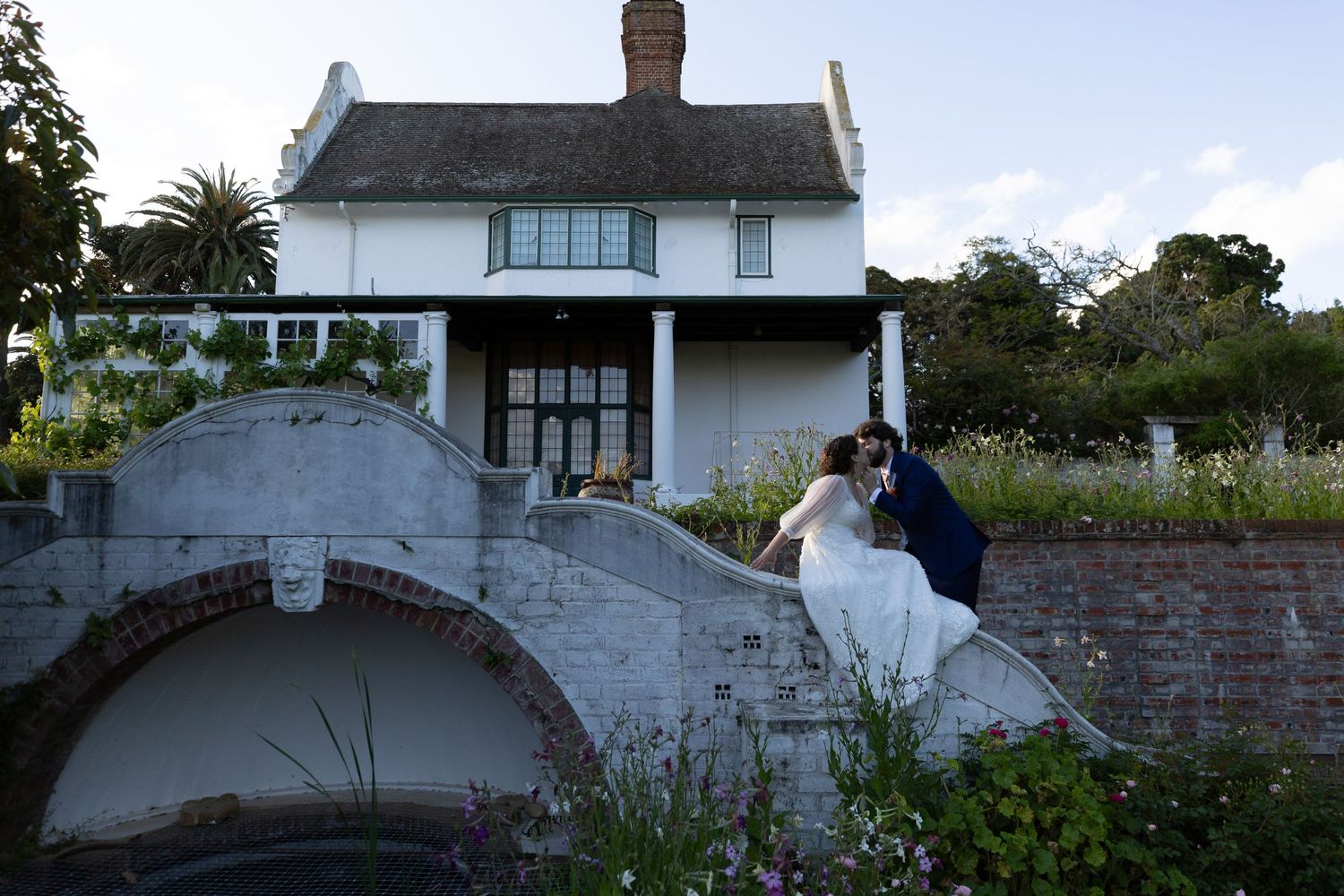 Bride and groom share a romantic kiss on a stone bridge outside a charming white cottage surrounded by lush gardens.