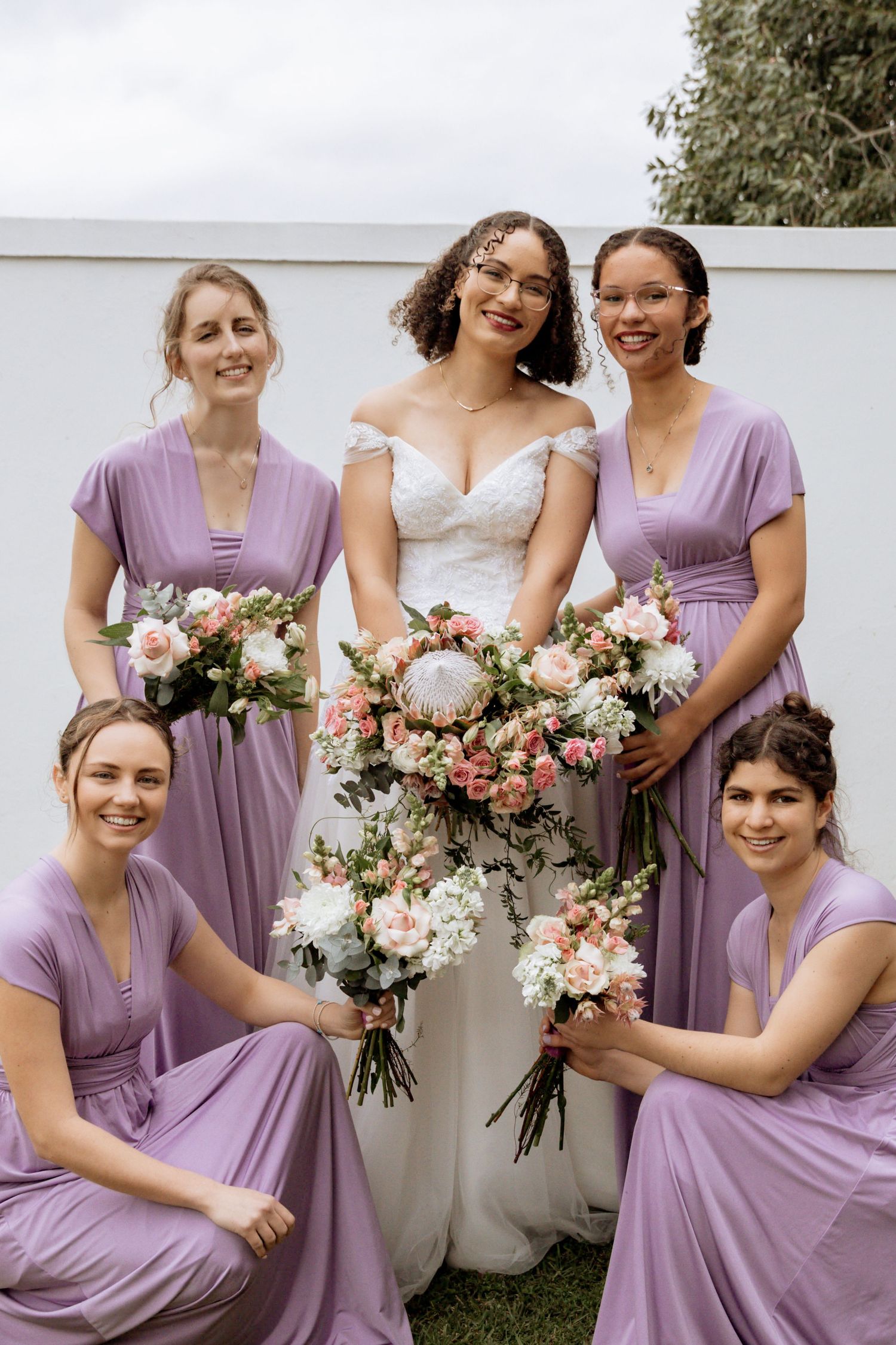 Wedding party in lavender bridesmaid dresses holding floral bouquets with pink and white flowers.