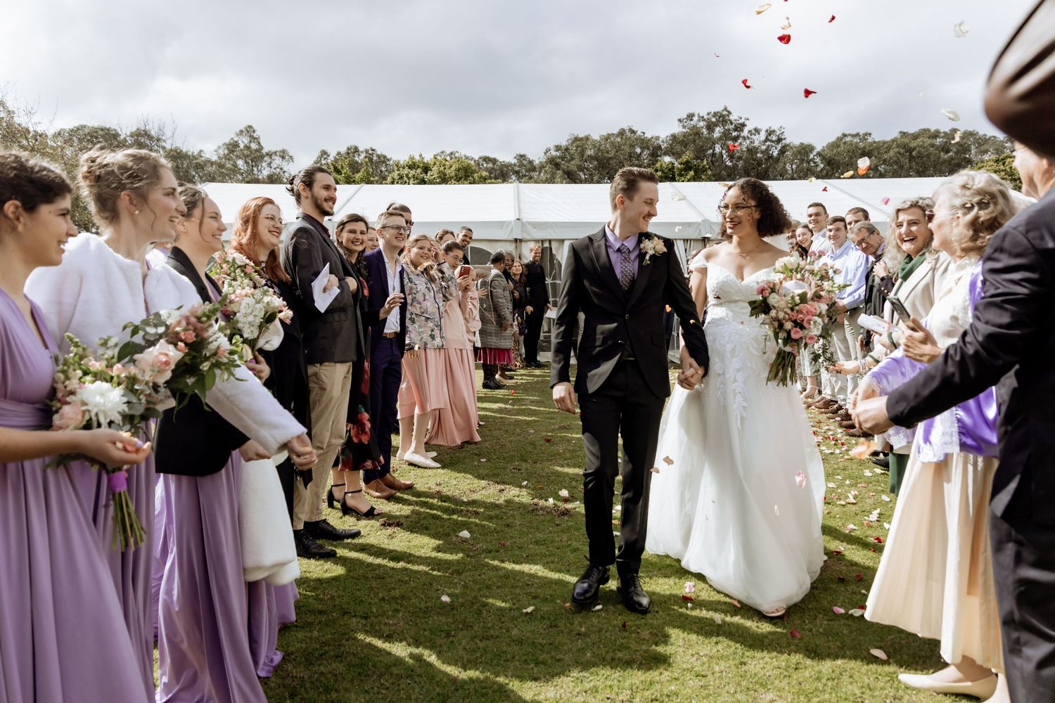 Wedding guests in lavender dresses line a grassy aisle while throwing flower petals during an outdoor ceremony.