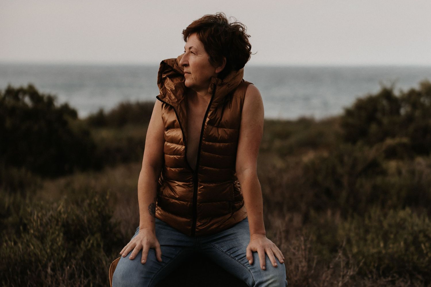 A person in a brown vest sits thoughtfully on coastal cliffs during golden hour as waves crash below.