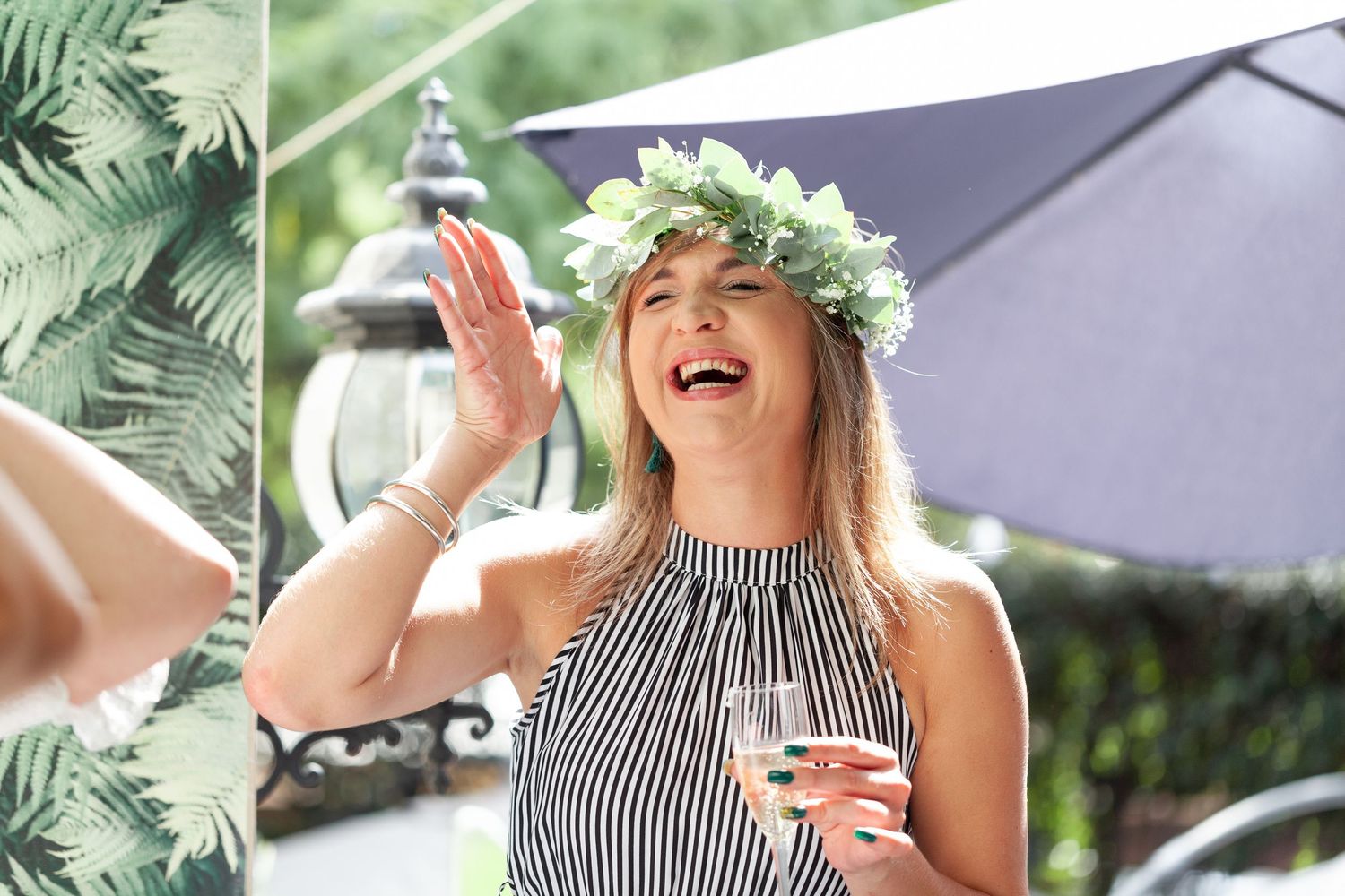 Laughing woman wearing floral crown and striped top holding wine glass at outdoor garden party.
