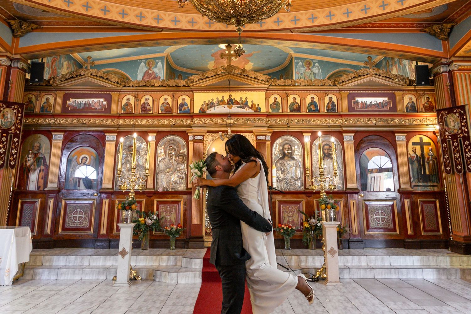 Bride and groom sharing a romantic kiss inside an ornate Orthodox church with golden iconostasis.