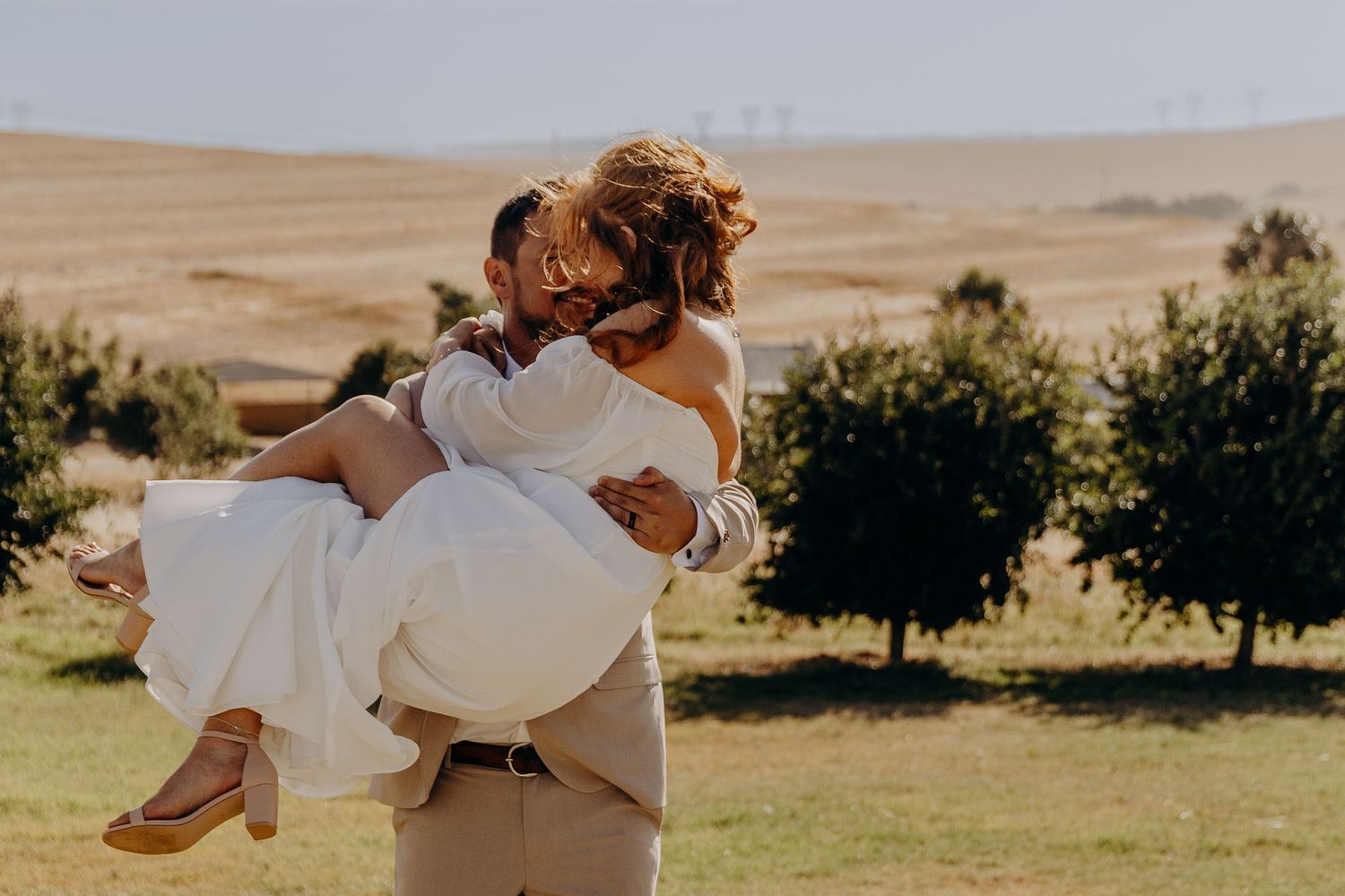 Groom in tan suit carries bride in white off-shoulder gown, laughing outdoors with golden hills and trees behind.