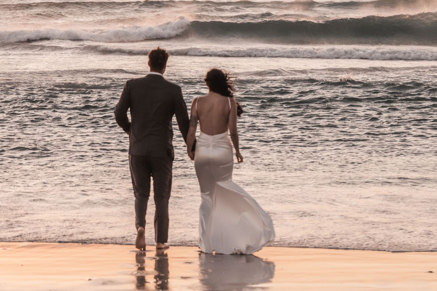 Newlywed couple in wedding attire walking hand in hand along a sandy beach with ocean waves behind them.
