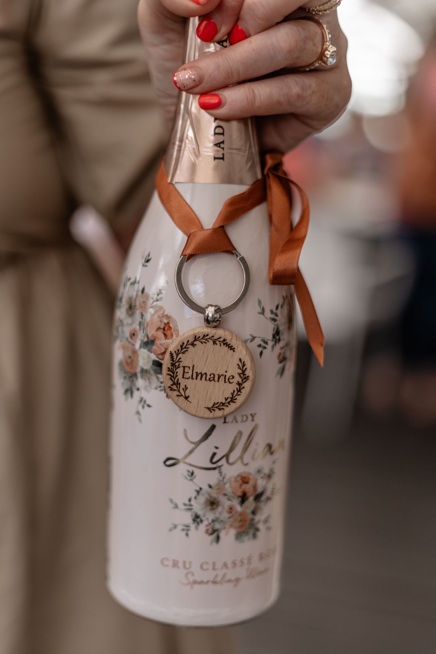 Hands with red nail polish holding a white wine bottle with elegant floral design and calligraphy lettering.
