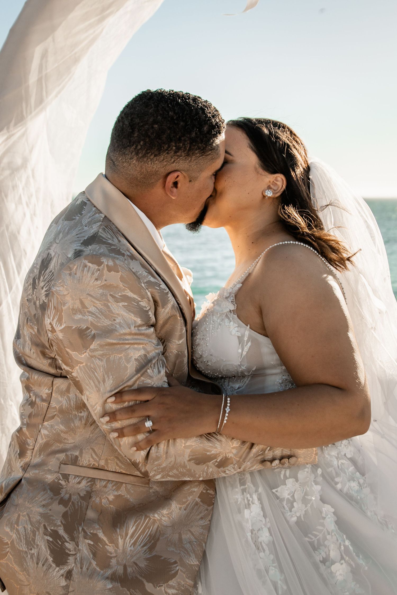 A romantic seaside wedding moment with a couple embracing and kissing in soft golden light.
