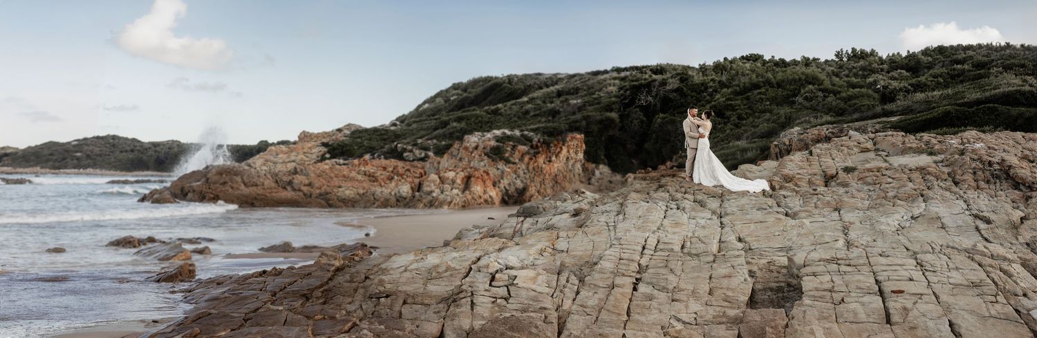 A couple stands at a scenic rocky coastal lookout with rugged cliffs and ocean waves in the background.
