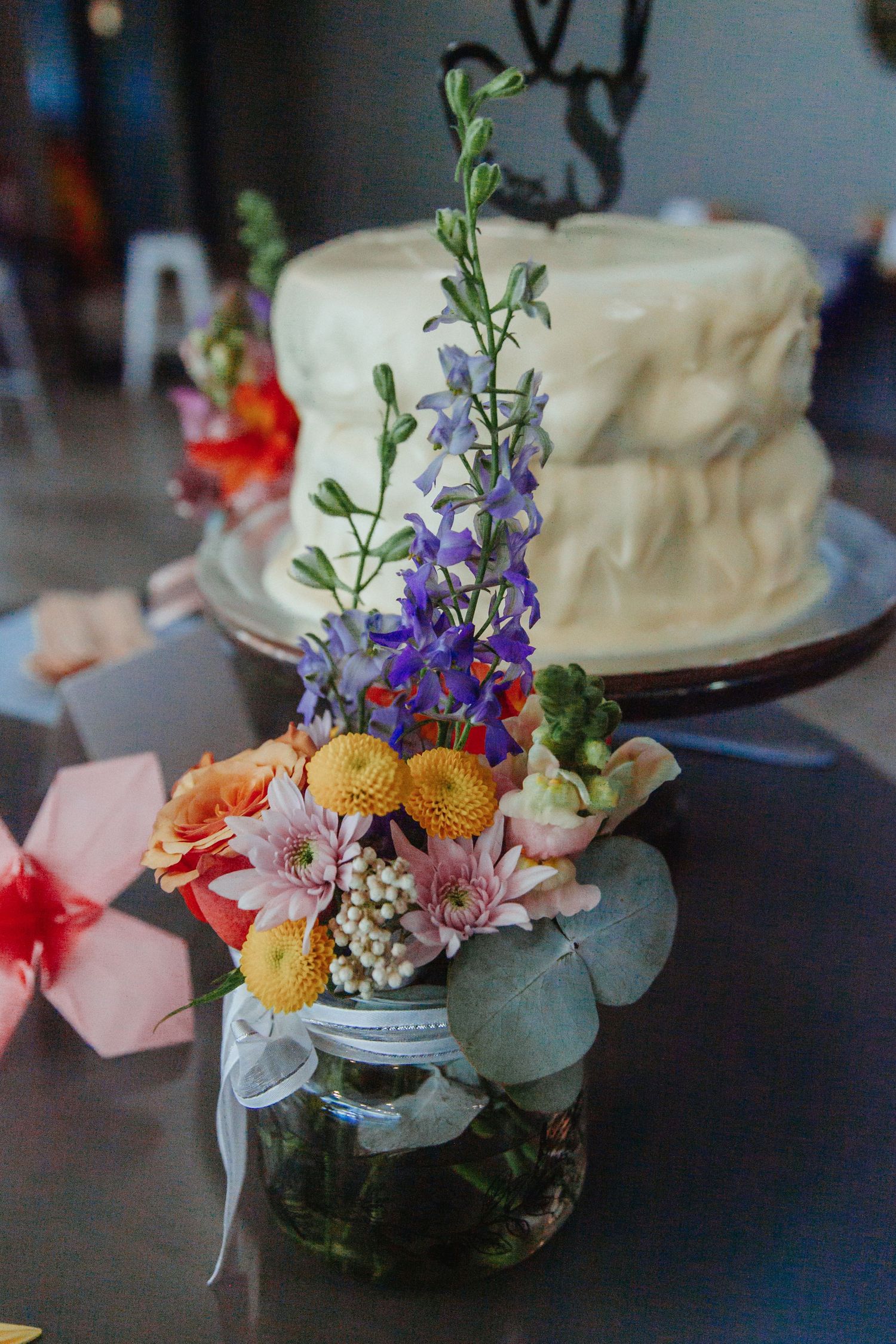 A rustic mason jar floral arrangement with purple delphinium and orange blooms sits beside a white frosted cake on display.