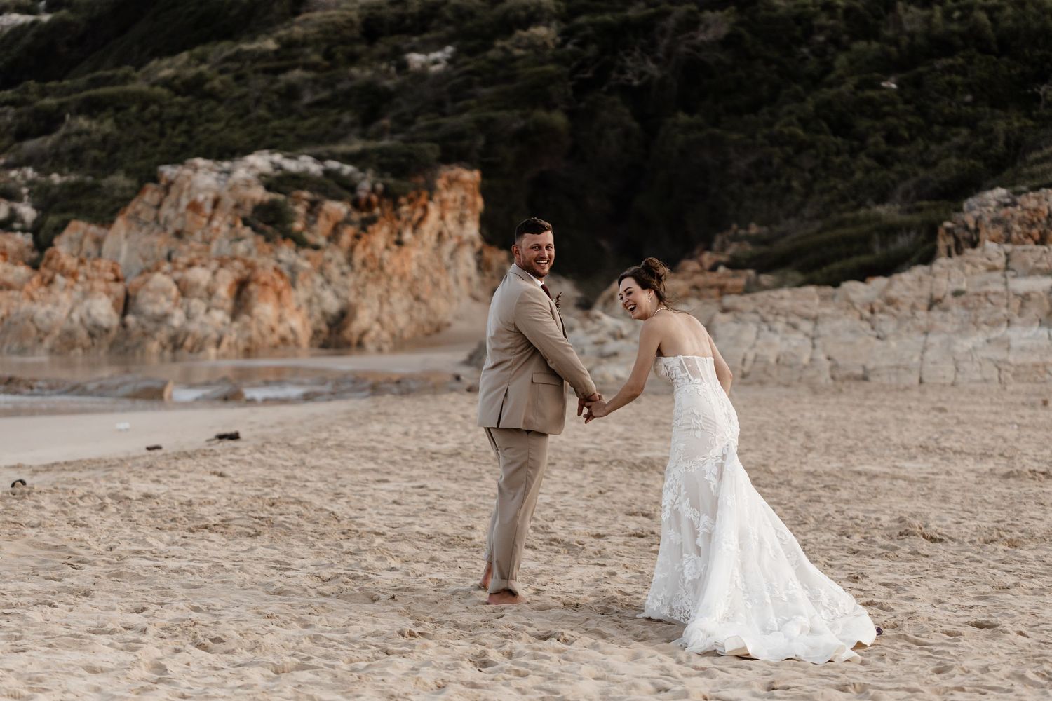 Couple walking along a scenic beach with rocky cliffs and natural landscape in the background.