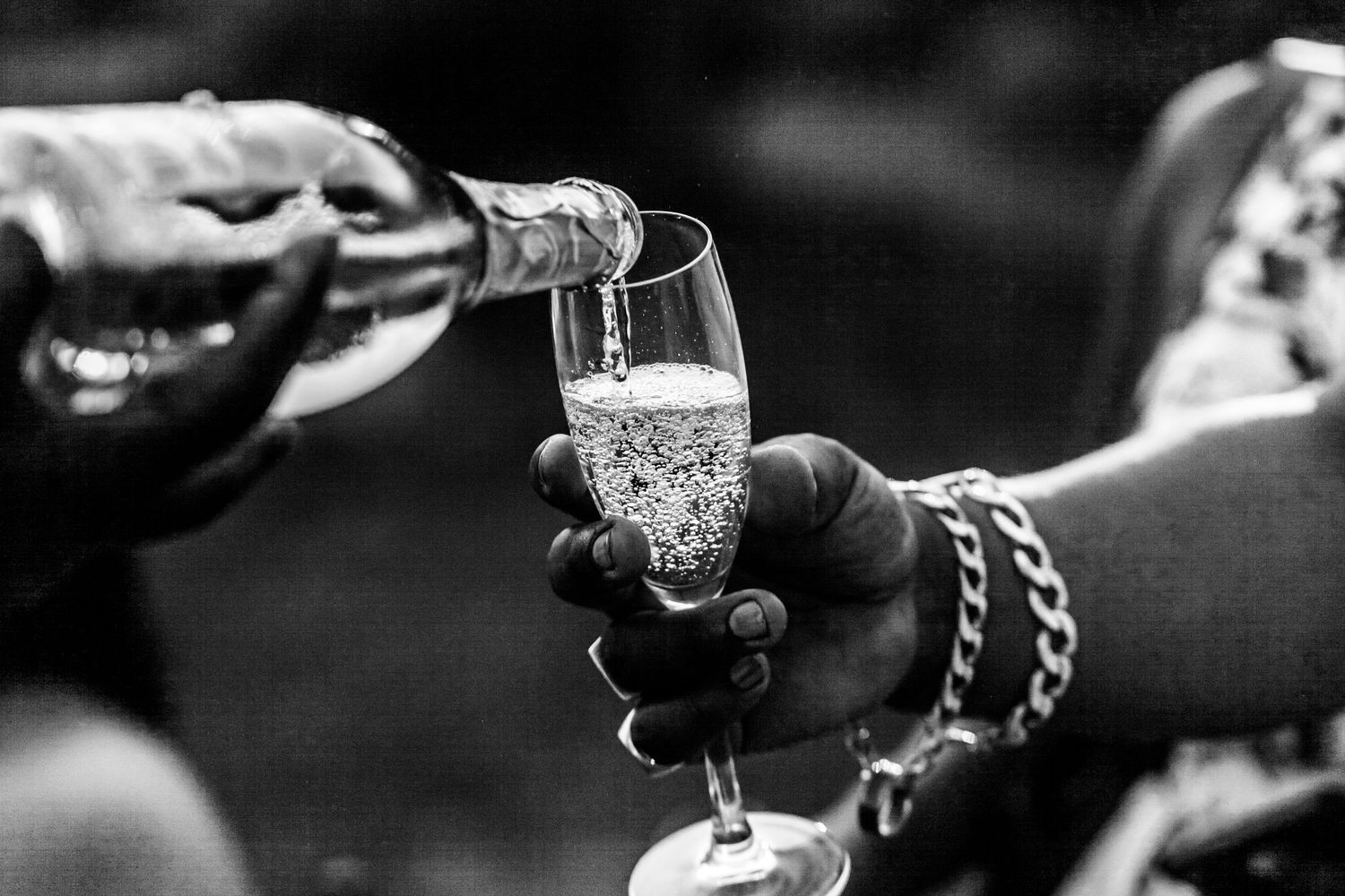 A hand pours champagne into a glass held by another hand wearing a bracelet in black and white photography.