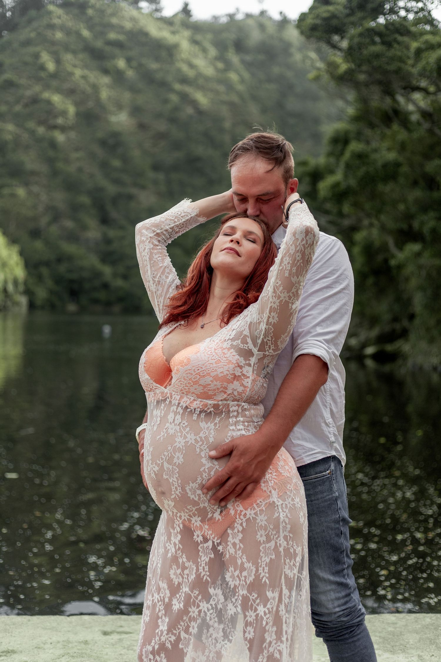 Romantic maternity photoshoot by a lake features couple in white lace dress and casual attire embracing outdoors.