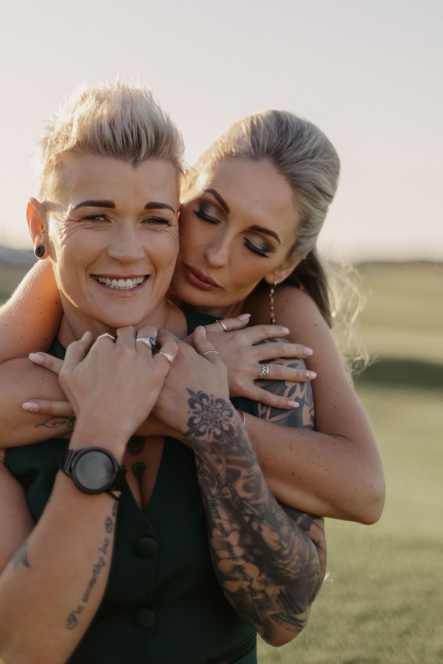 Two people with tattoos share intimate candid moments while laughing together in a grassy field.
