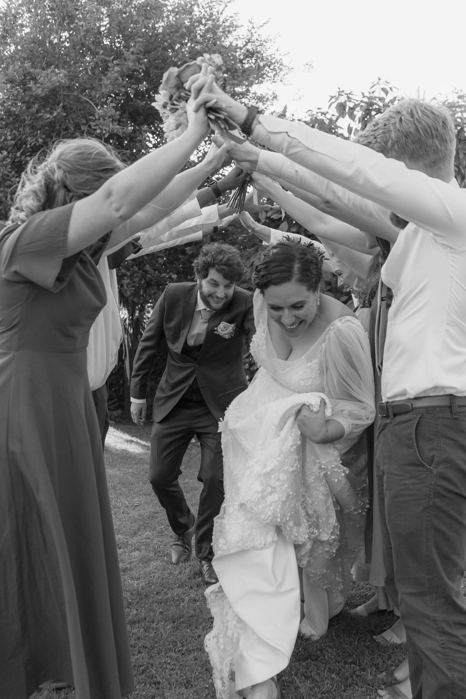 Wedding party members form a tunnel arch with their arms while a couple in formal attire ducks underneath during celebrations.