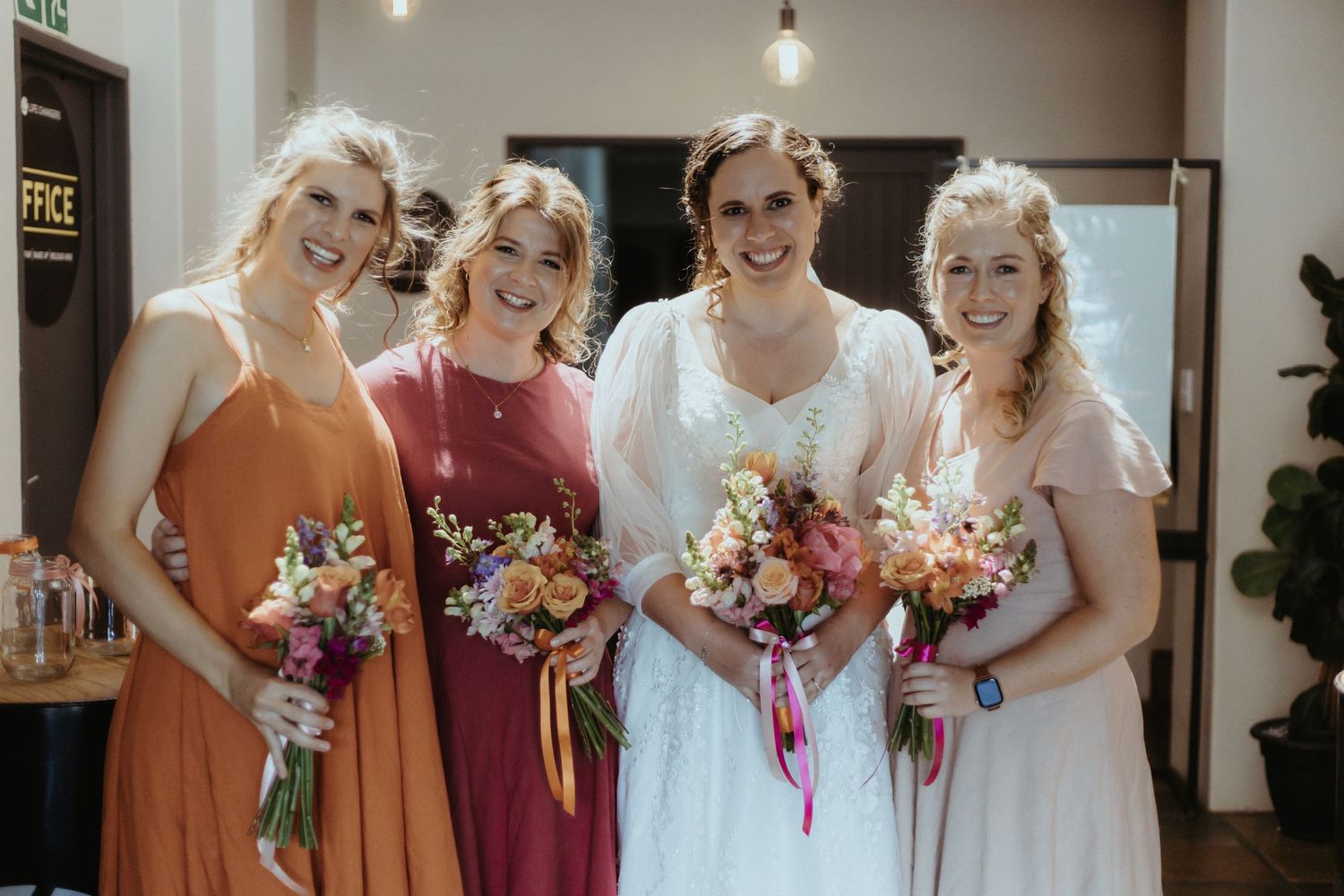 A bridal party in orange, burgundy and white dresses hold colorful bouquets while smiling indoors.