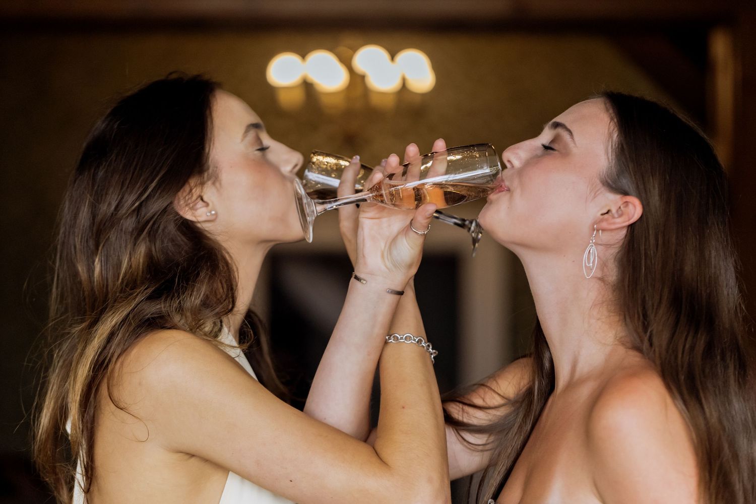 Two bridesmaids sharing a champagne toast, drinking from the same glass at a wedding celebration.