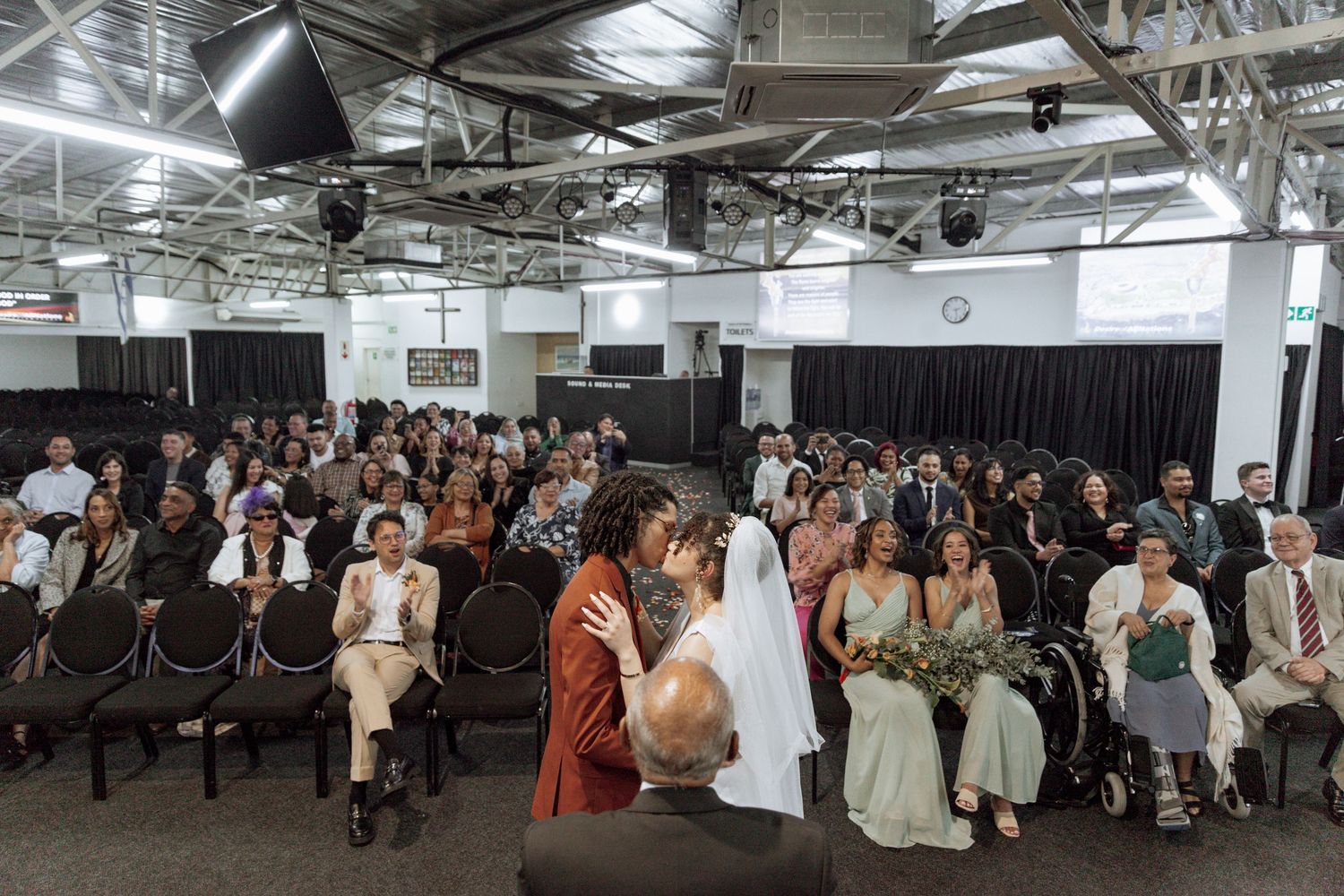 Wedding guests seated in rows of black chairs at an industrial-style venue with exposed ceiling and dark curtains.