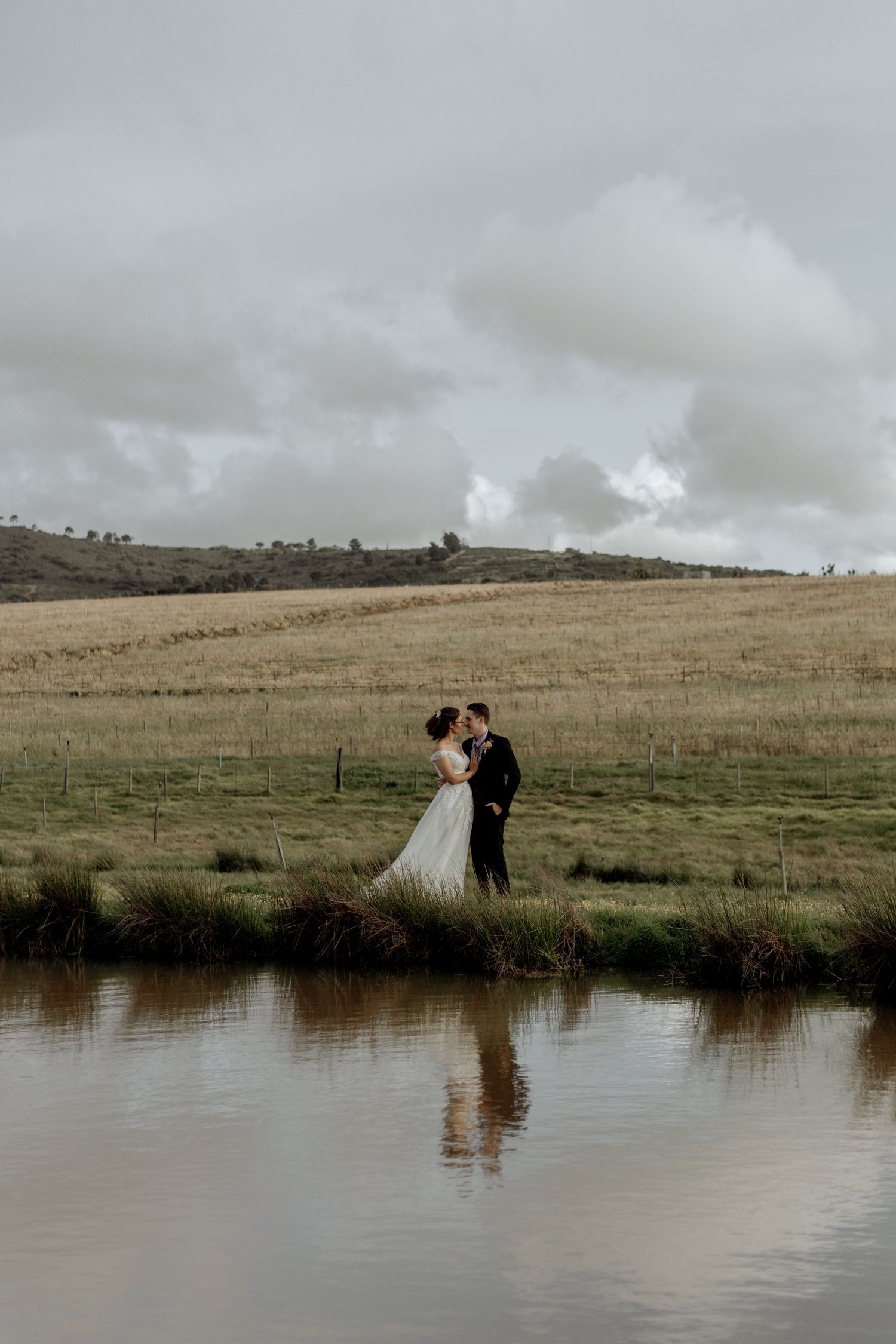Couple in wedding attire standing by a serene pond with cloudy skies and rolling hills in the background.