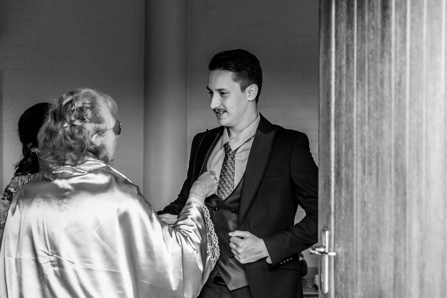 A black and white photograph shows two people in conversation near a door, with one wearing a suit and the other in satin.