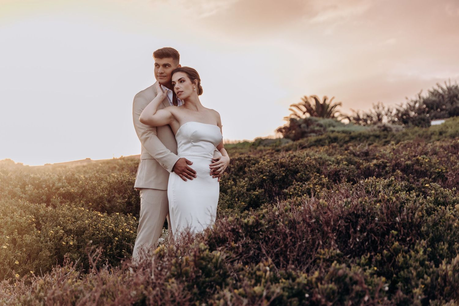 Bride and groom embracing at sunset in a field of wildflowers, wearing elegant white dress and beige suit.