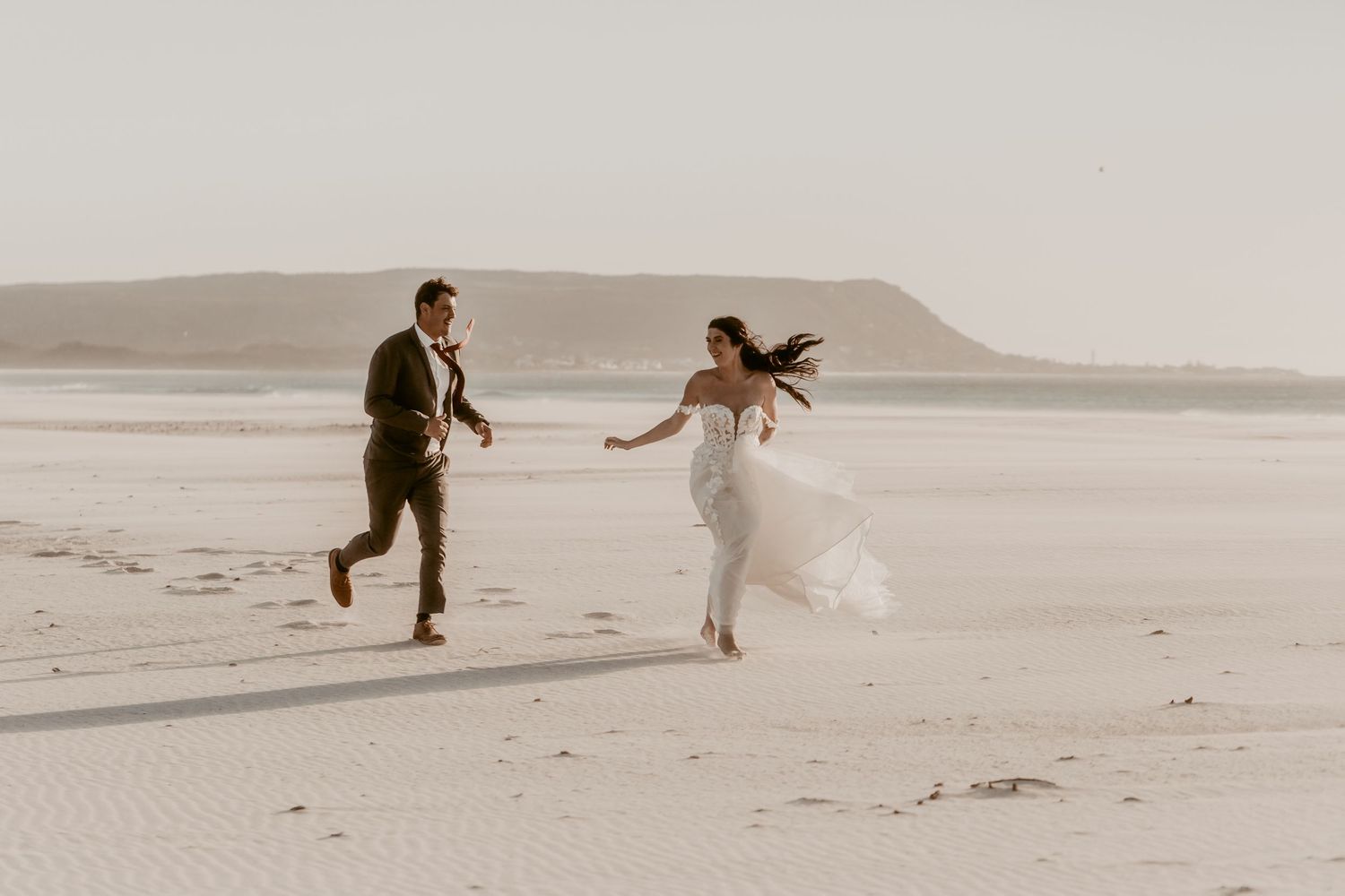 Bride and groom running joyfully on a sandy beach in soft sunlight, bride in white gown, groom in dark suit.