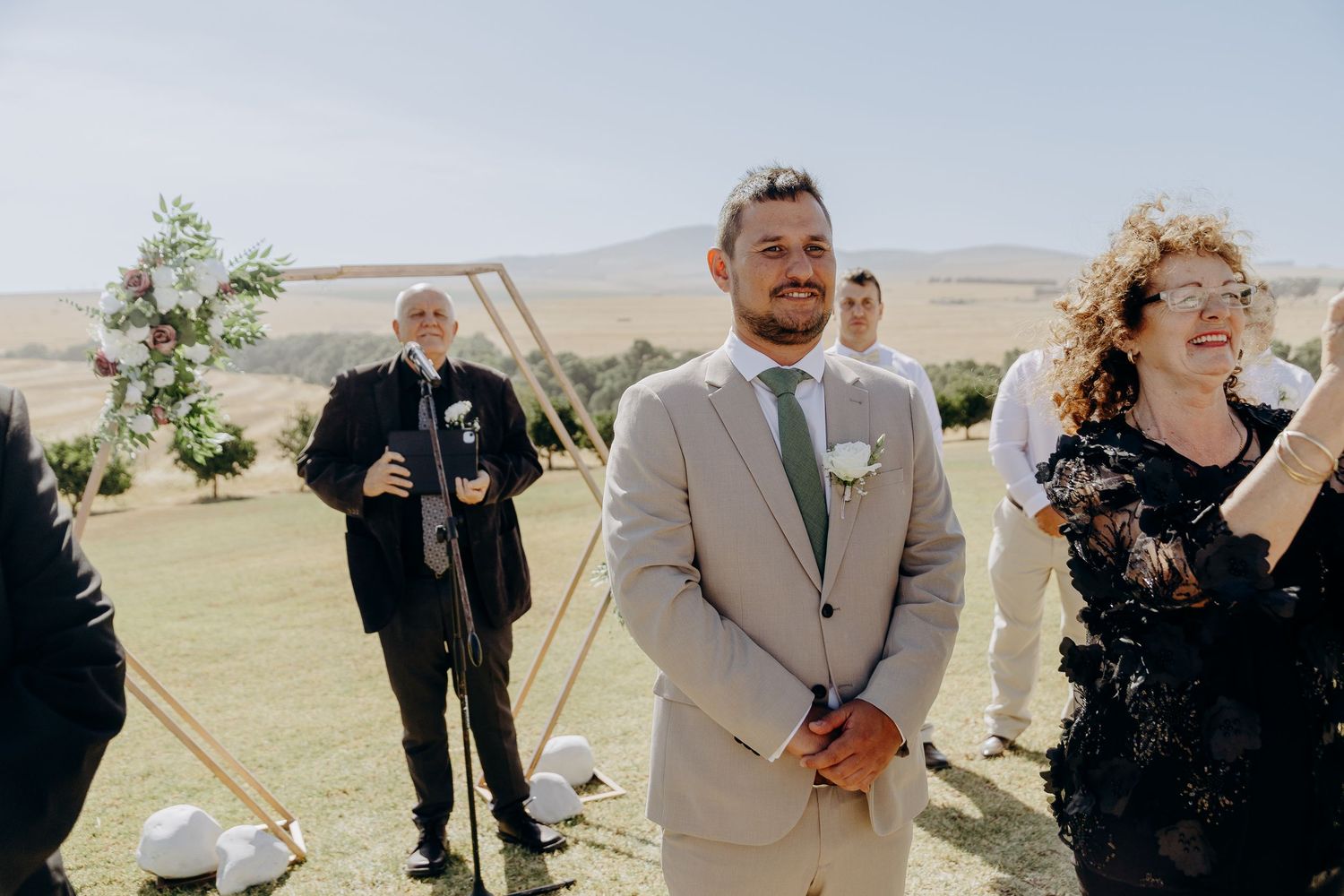 Groom in beige suit smiling at outdoor wedding ceremony on green hillside with guests and floral arch backdrop.