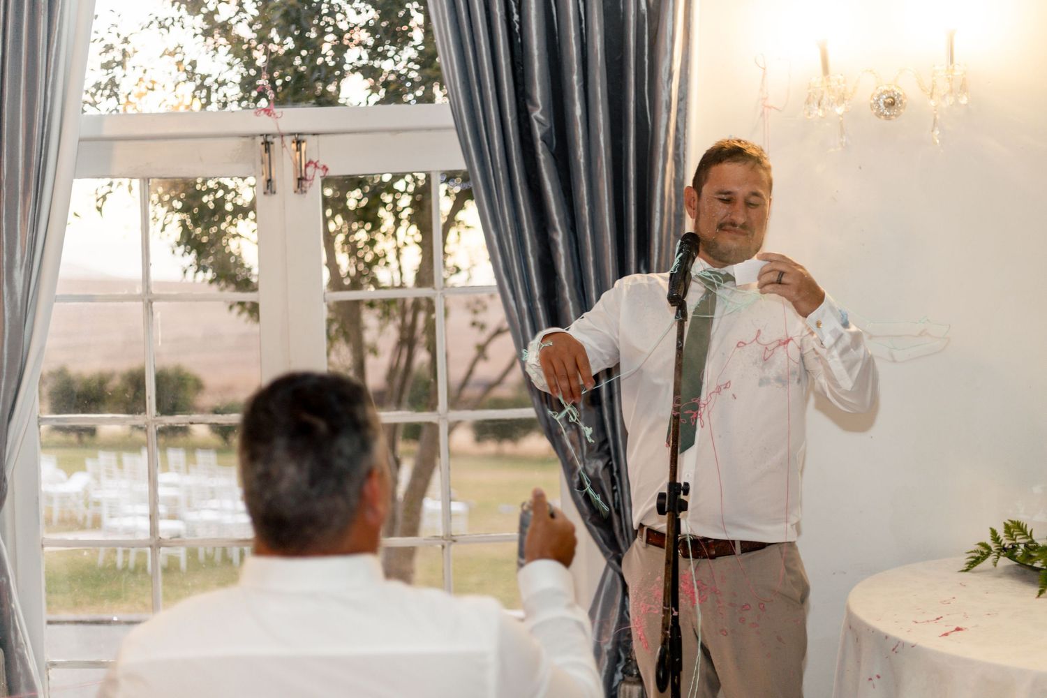 Man in white shirt giving a speech at an elegant wedding reception near large windows.