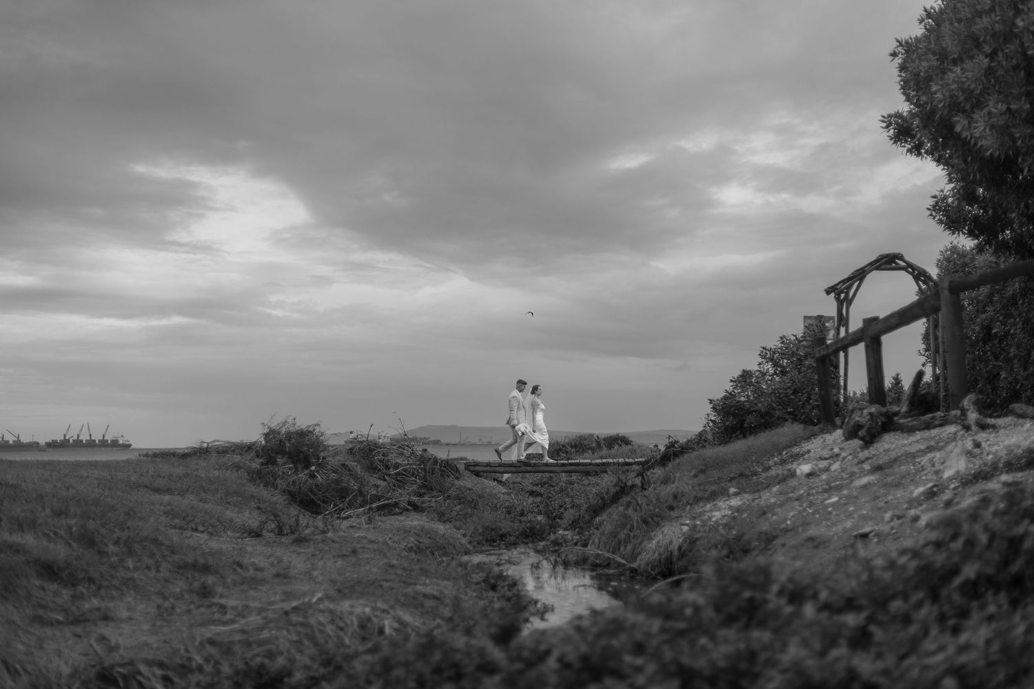 Black and white photo of a couple embracing on a grassy hillside under dramatic cloudy skies near a rustic arbor.