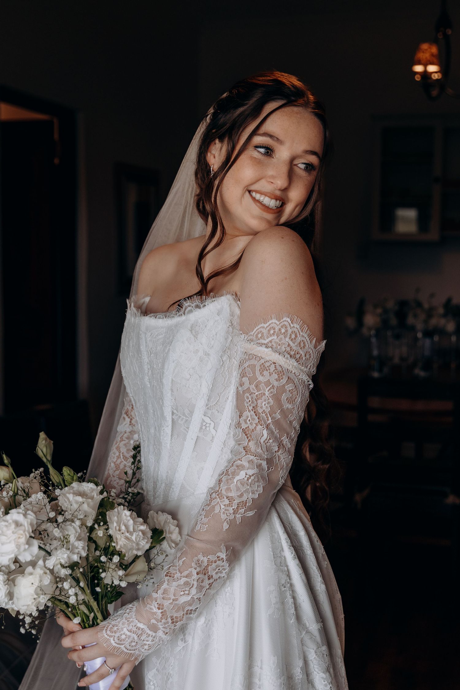 Smiling bride in off-shoulder lace wedding dress with veil holding white floral bouquet indoors.