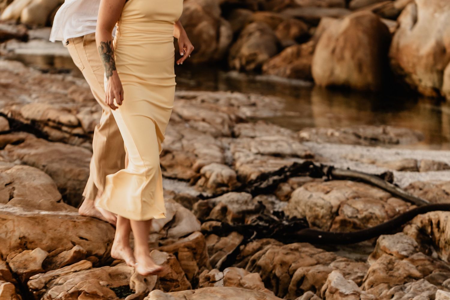 Two people walk along rocky coastal terrain while wearing light beige colored clothing during sunset.