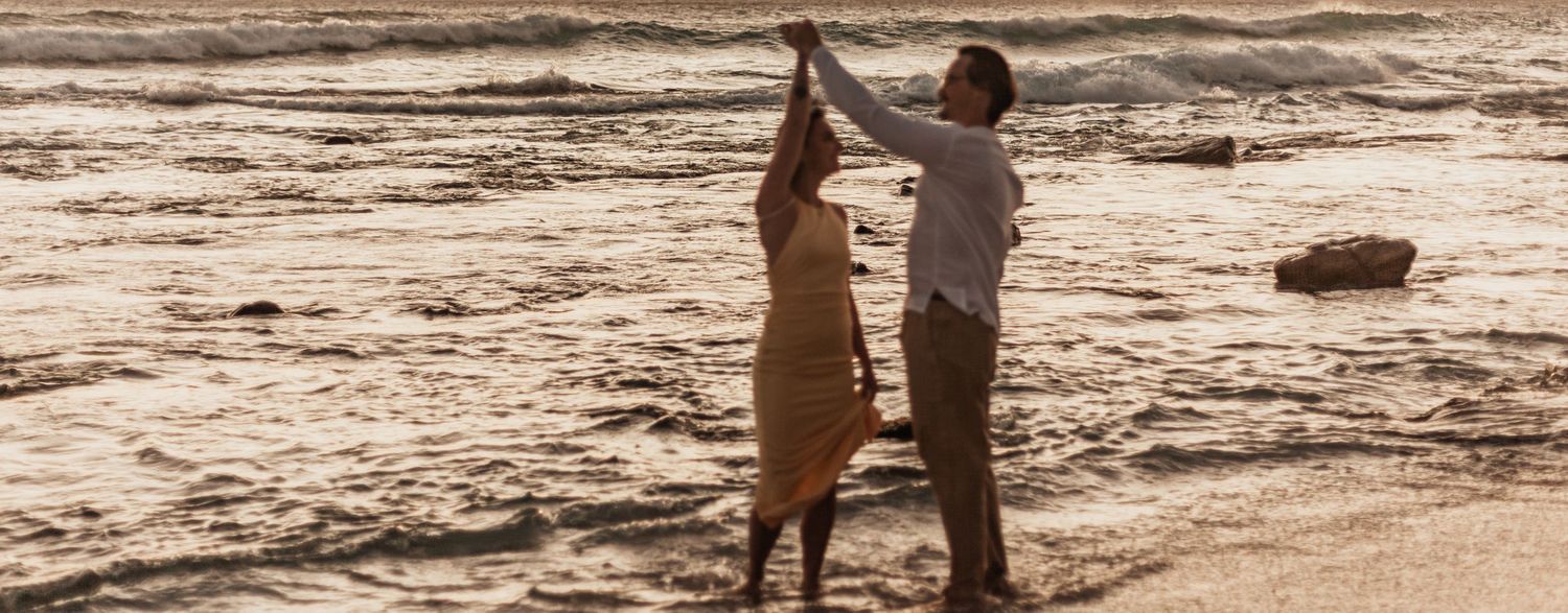 A silhouetted couple dances together at sunset by the ocean shore as waves roll in.