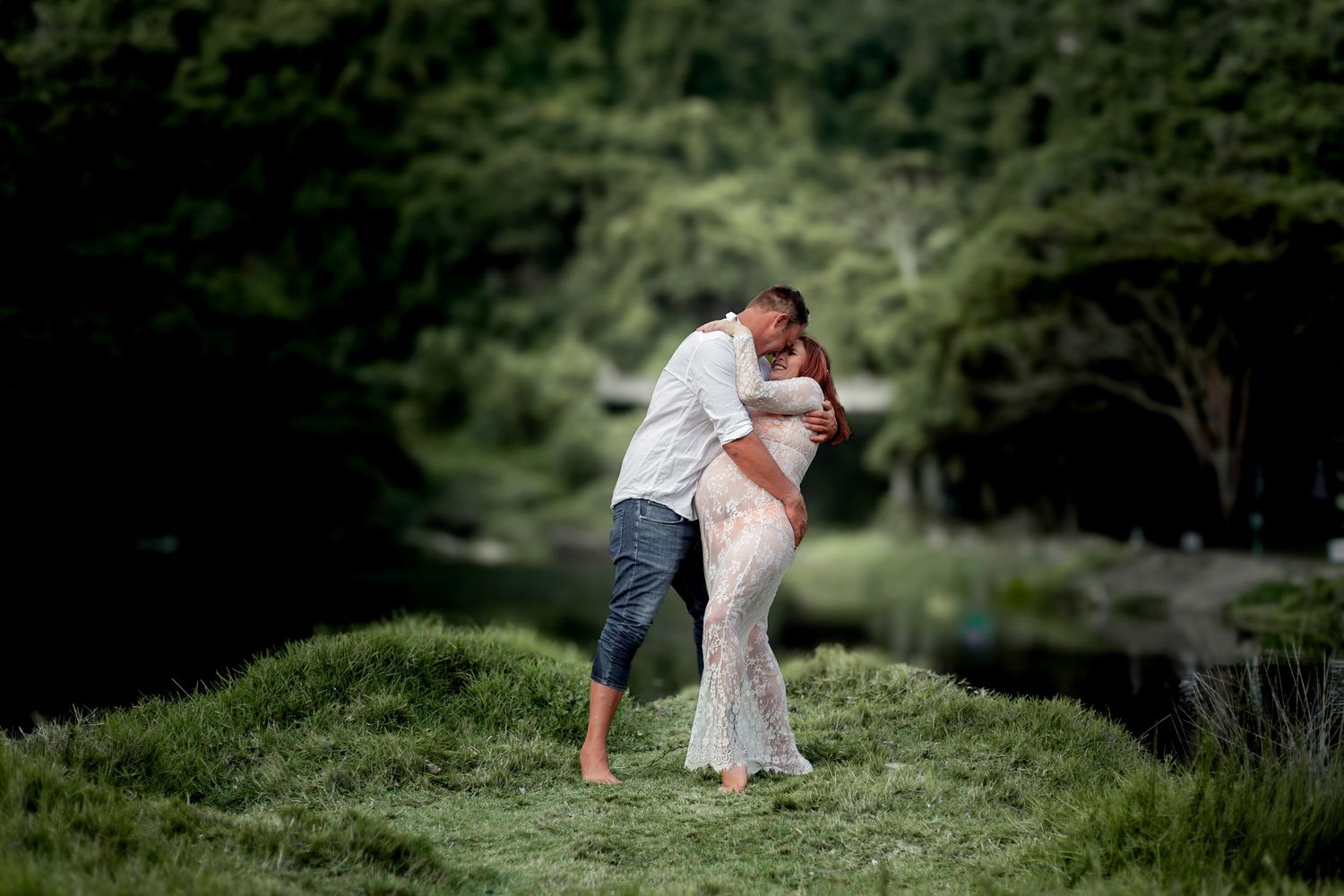 A couple embraces intimately on a grassy hillside with lush green forest in the background during a sunny day.