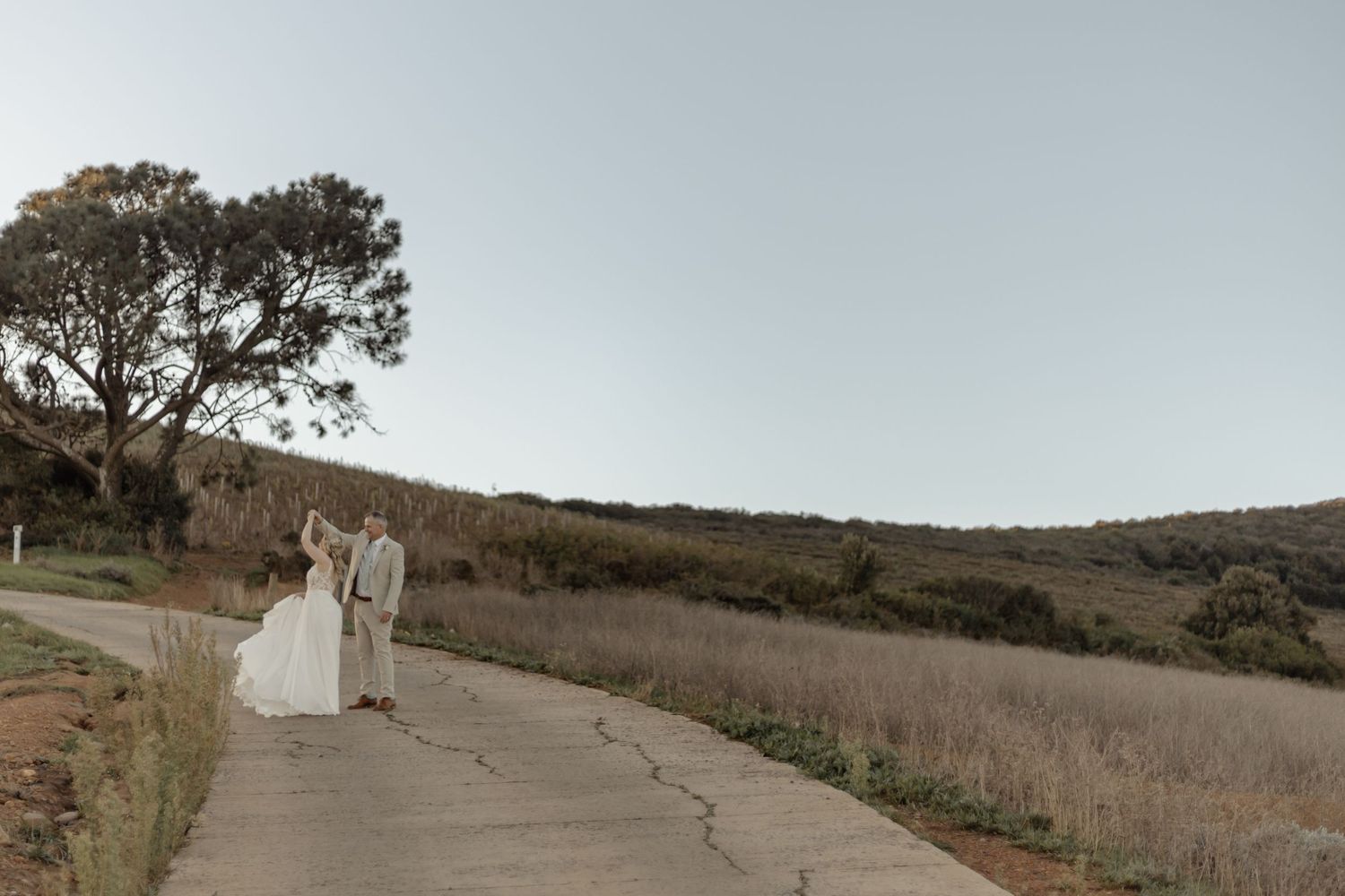 A couple in white wedding attire stands together on a scenic hillside path during golden hour.