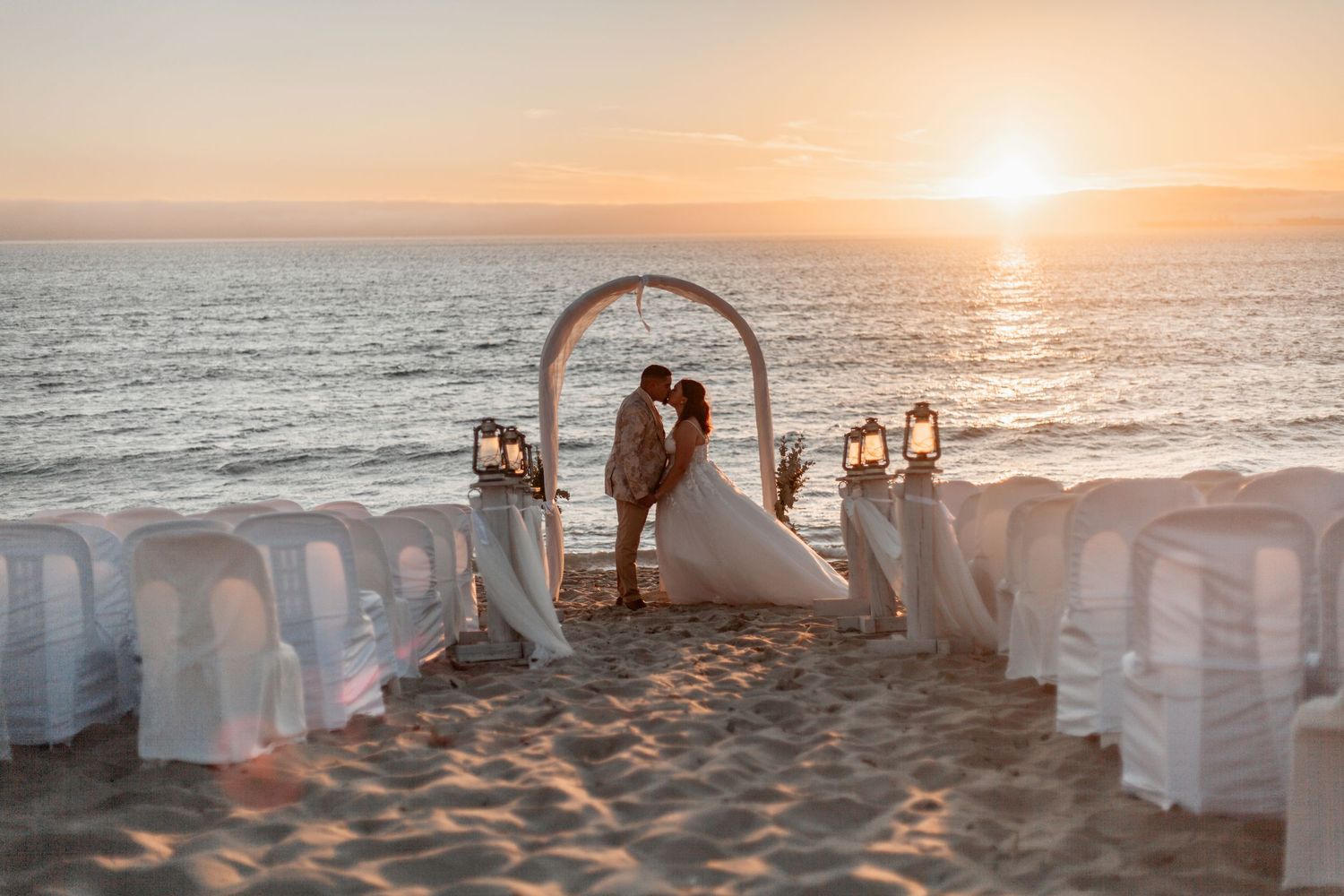 A romantic beach wedding ceremony setup with white chairs and floral arch at sunset over the ocean.