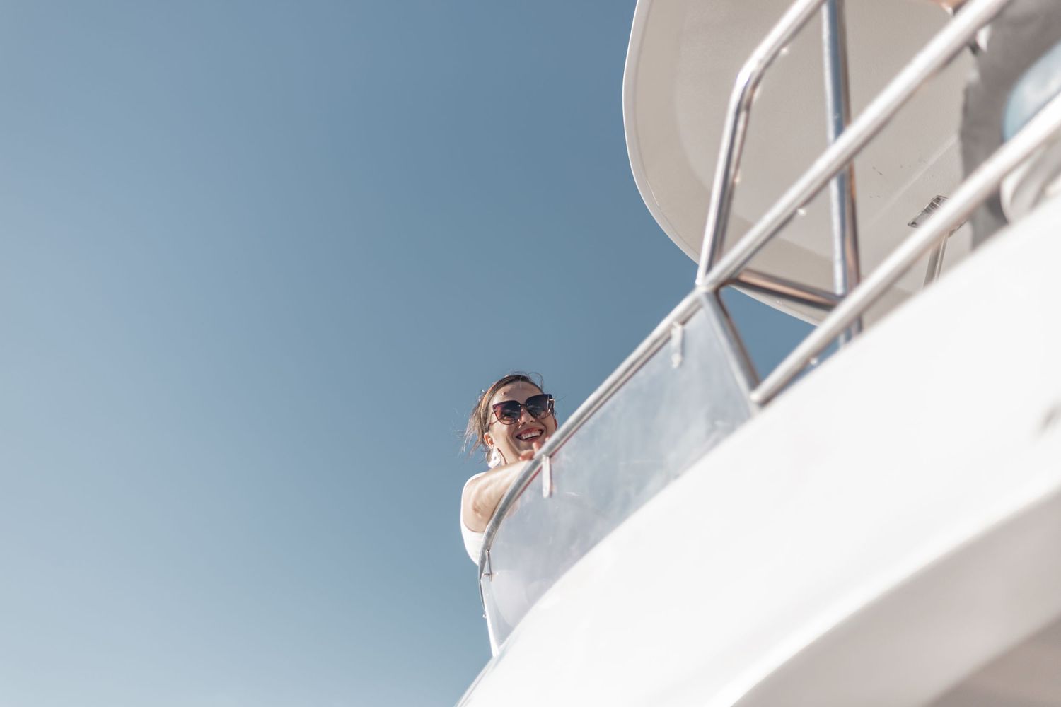 A person in sunglasses stands on the deck of a white yacht against a clear blue sky, looking out over the water.