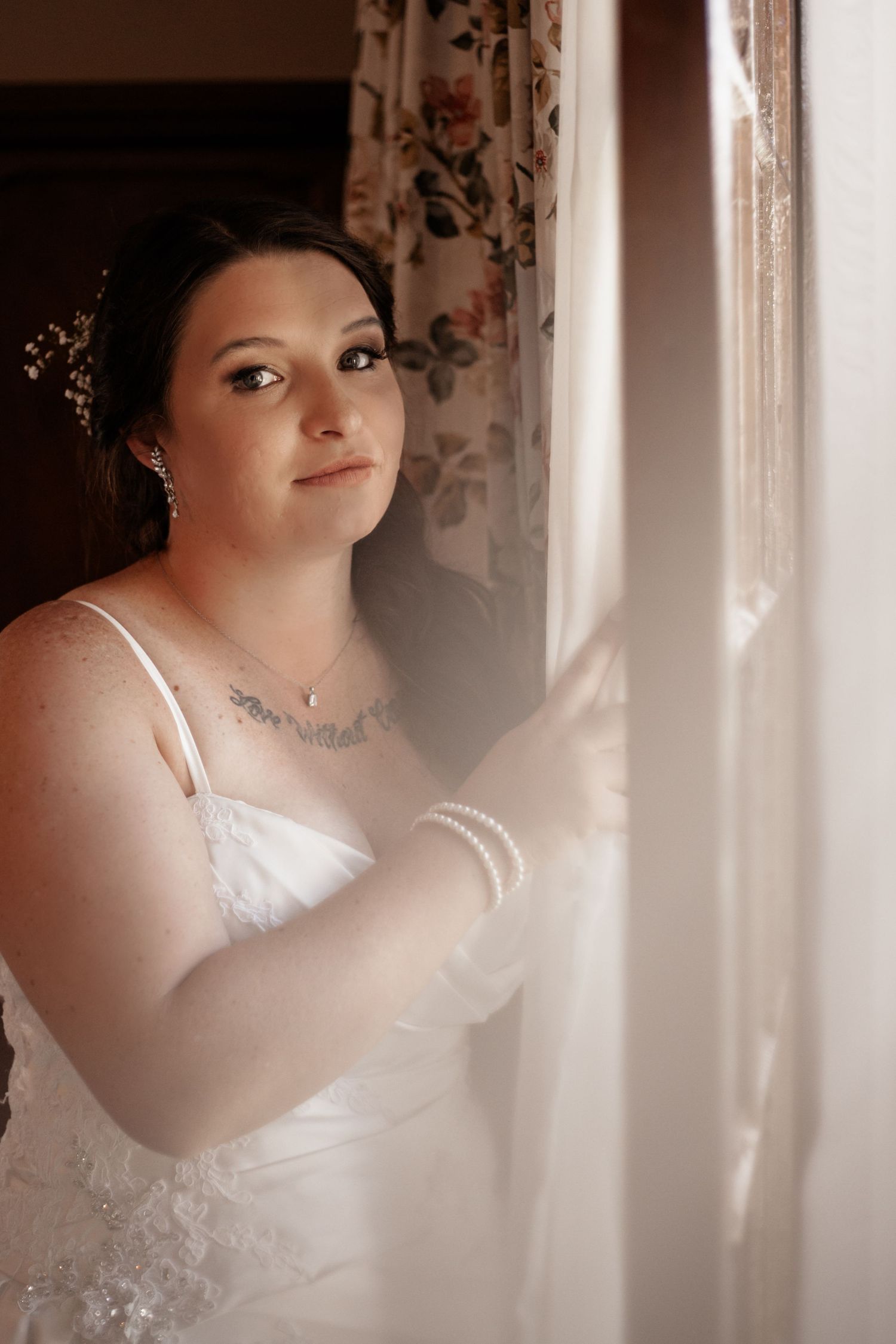 A bride in a white dress stands near a window in soft lighting with floral curtains in the background.