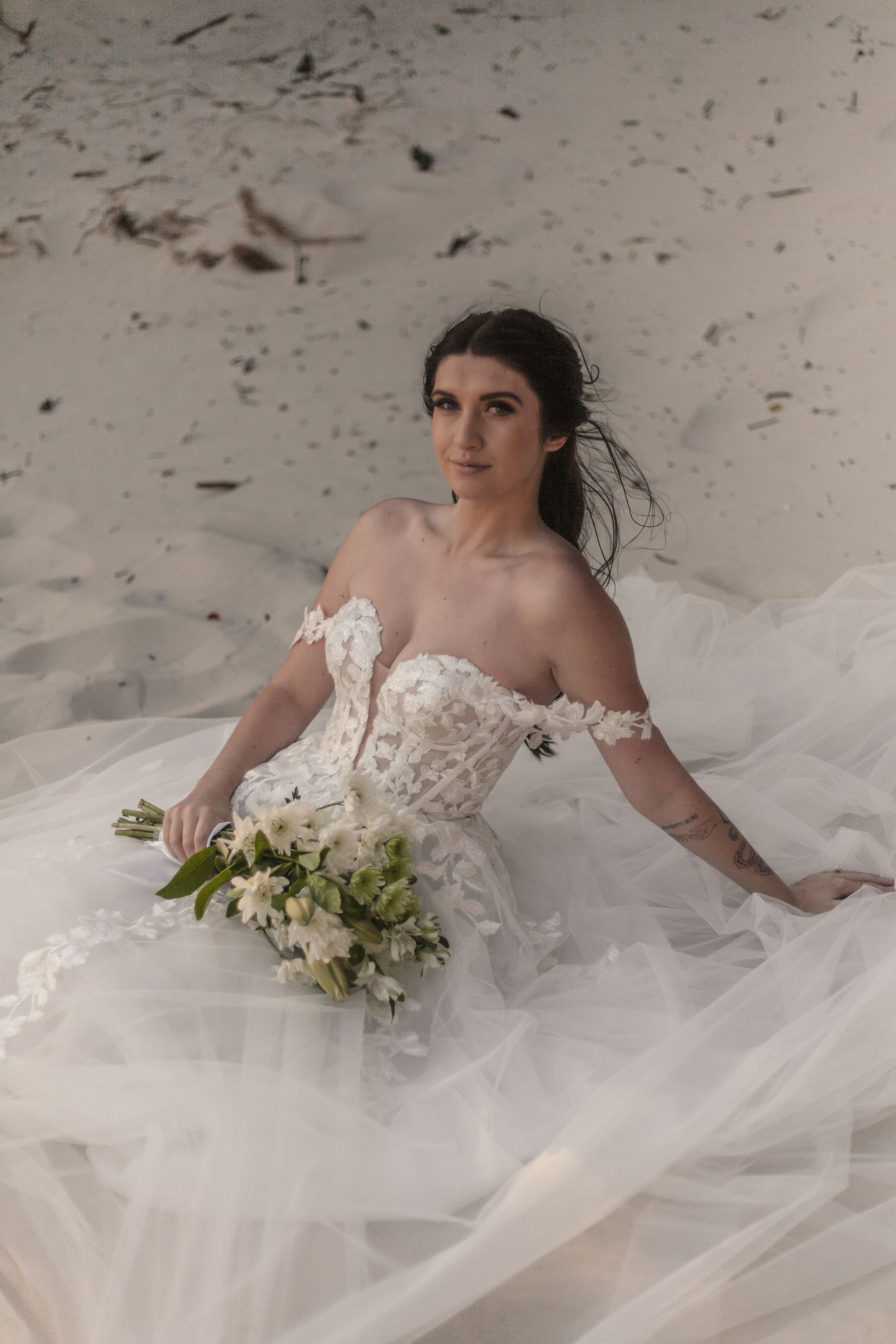 Bride in off-shoulder lace wedding gown sits on white sand holding white floral bouquet, gazing elegantly into distance.