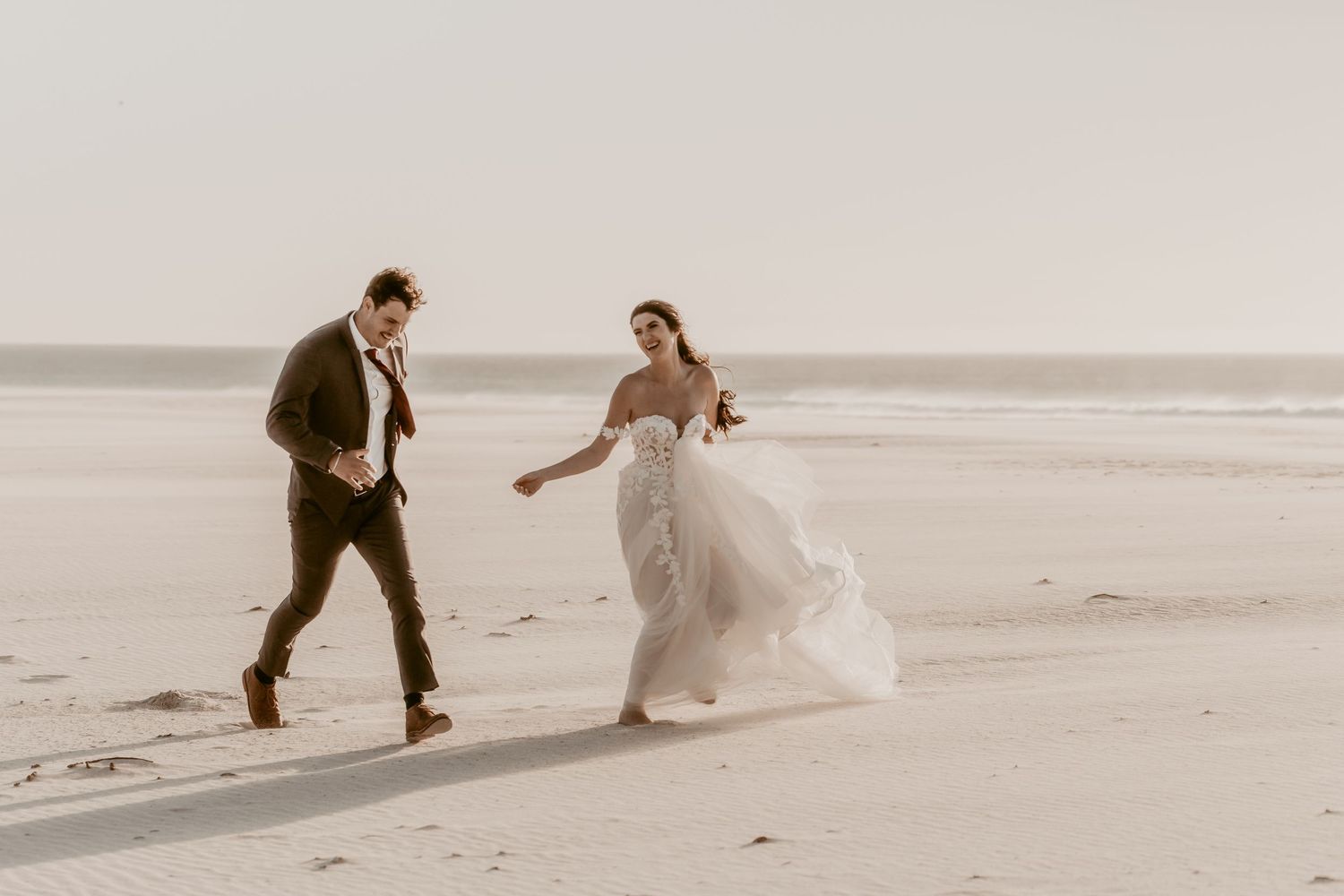 Bride and groom running joyfully on a sandy beach, bride in flowing white gown holding bouquet, groom in dark suit.