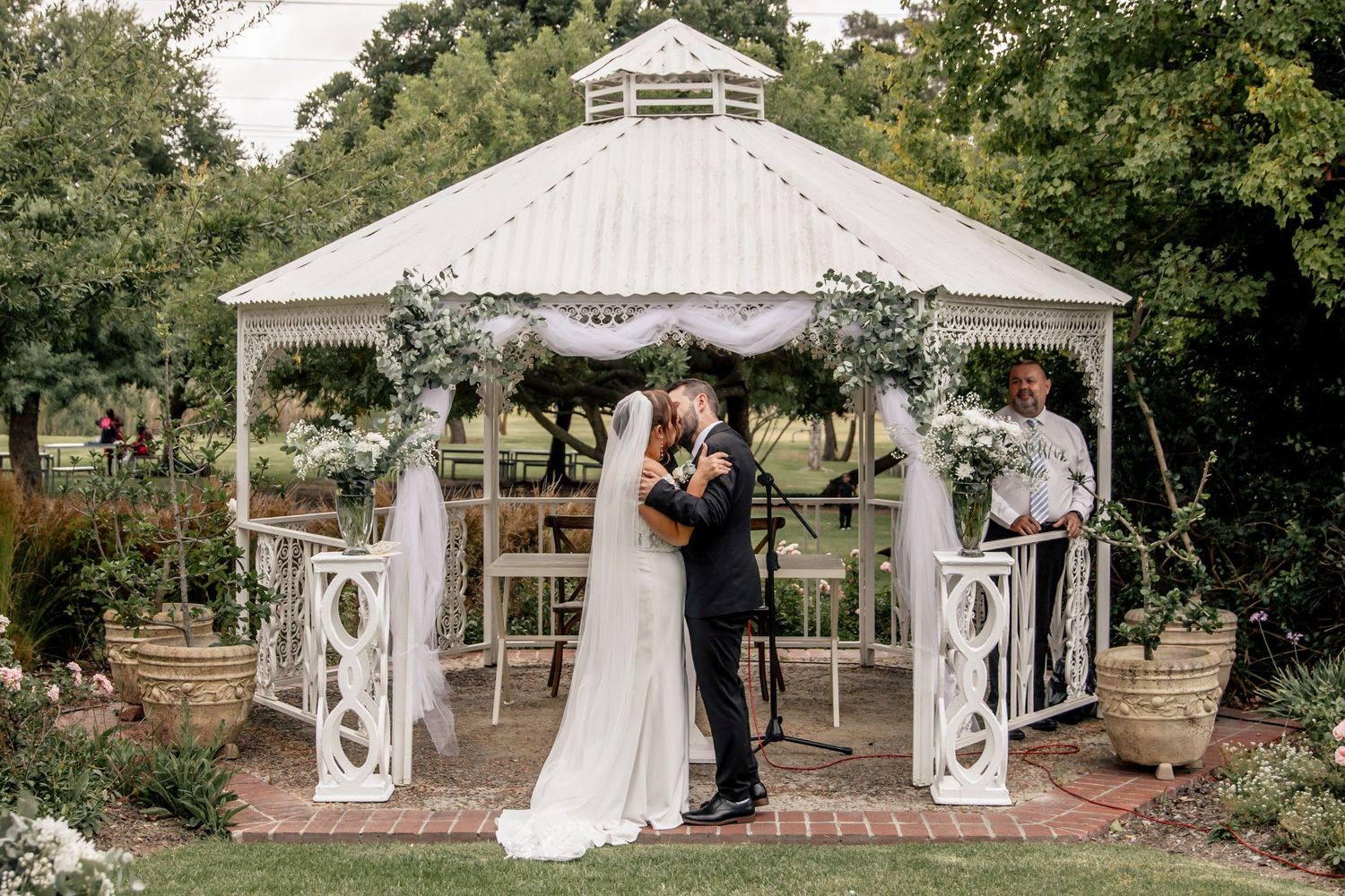 Bride and groom share first kiss under white gazebo adorned with floral arrangements in a lush garden wedding ceremony.
