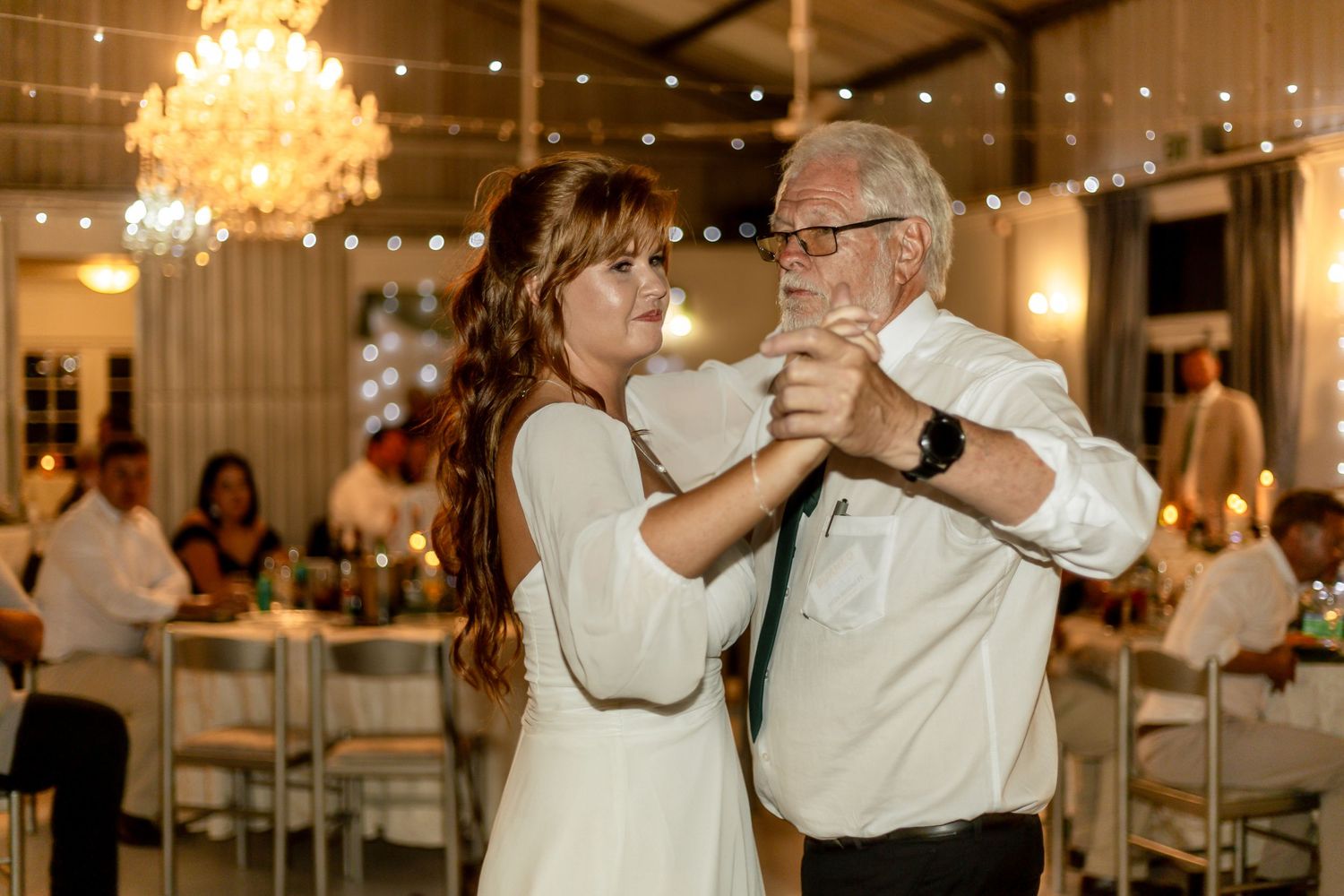 Bride and older man share a father-daughter dance at elegant wedding reception with string lights and chandelier.