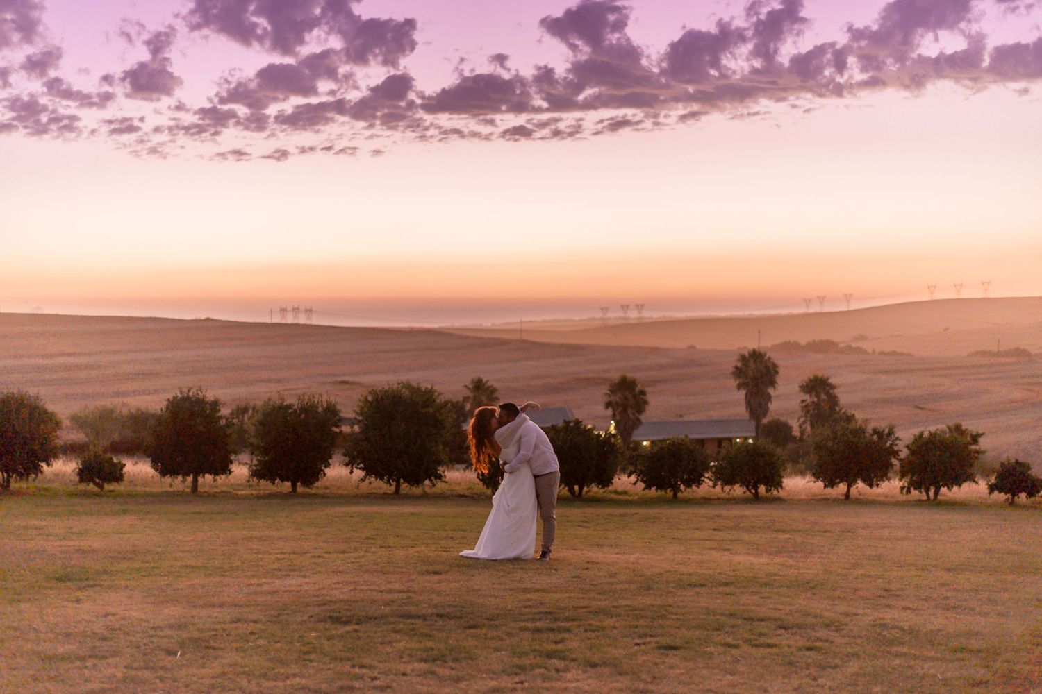 Couple sharing romantic embrace at sunset on green field with rolling hills, trees, and wind turbines in golden sky backdrop.