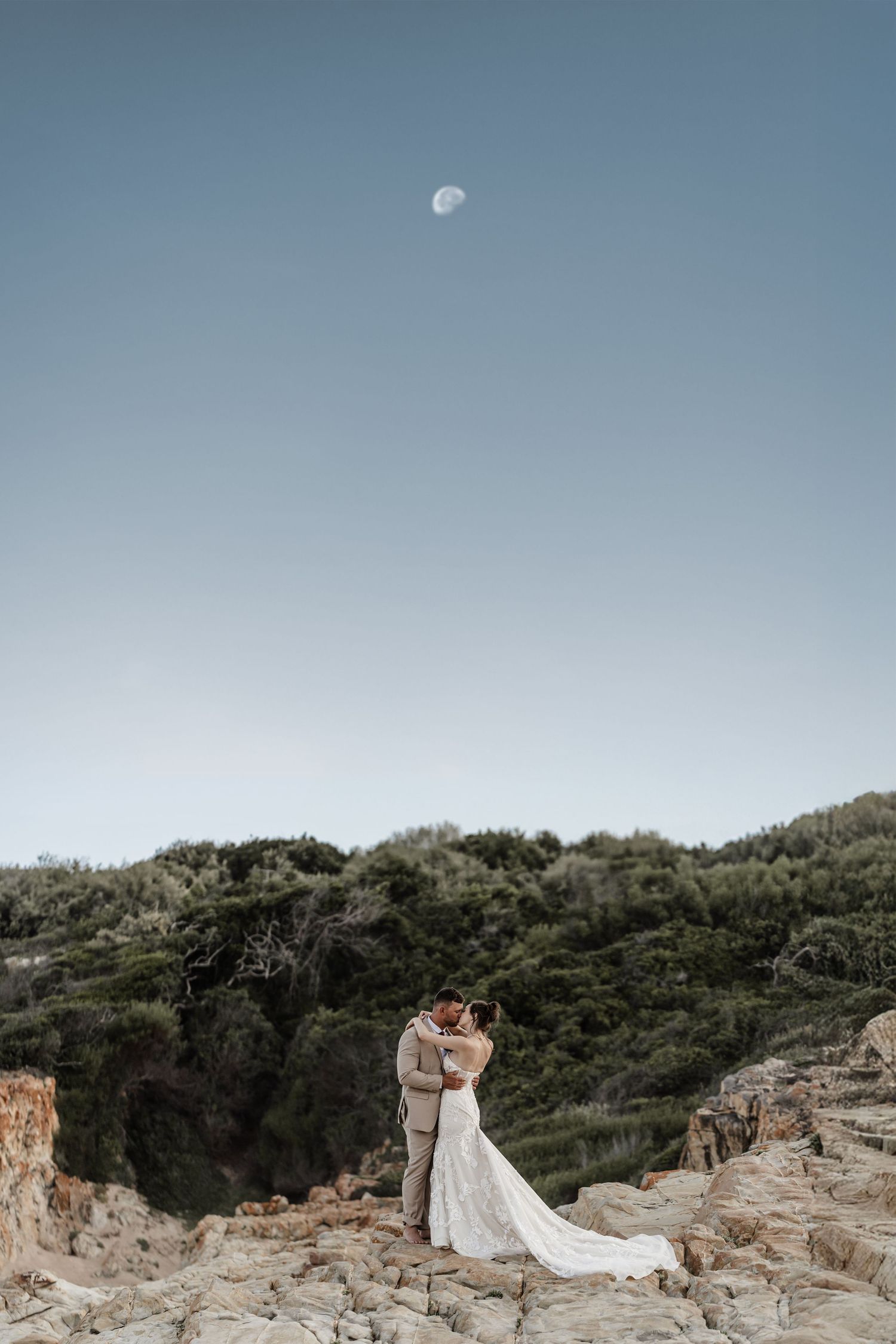 A panoramic view of a rocky coastal landscape with a moon in the sky and a white wedding dress on the cliffside.