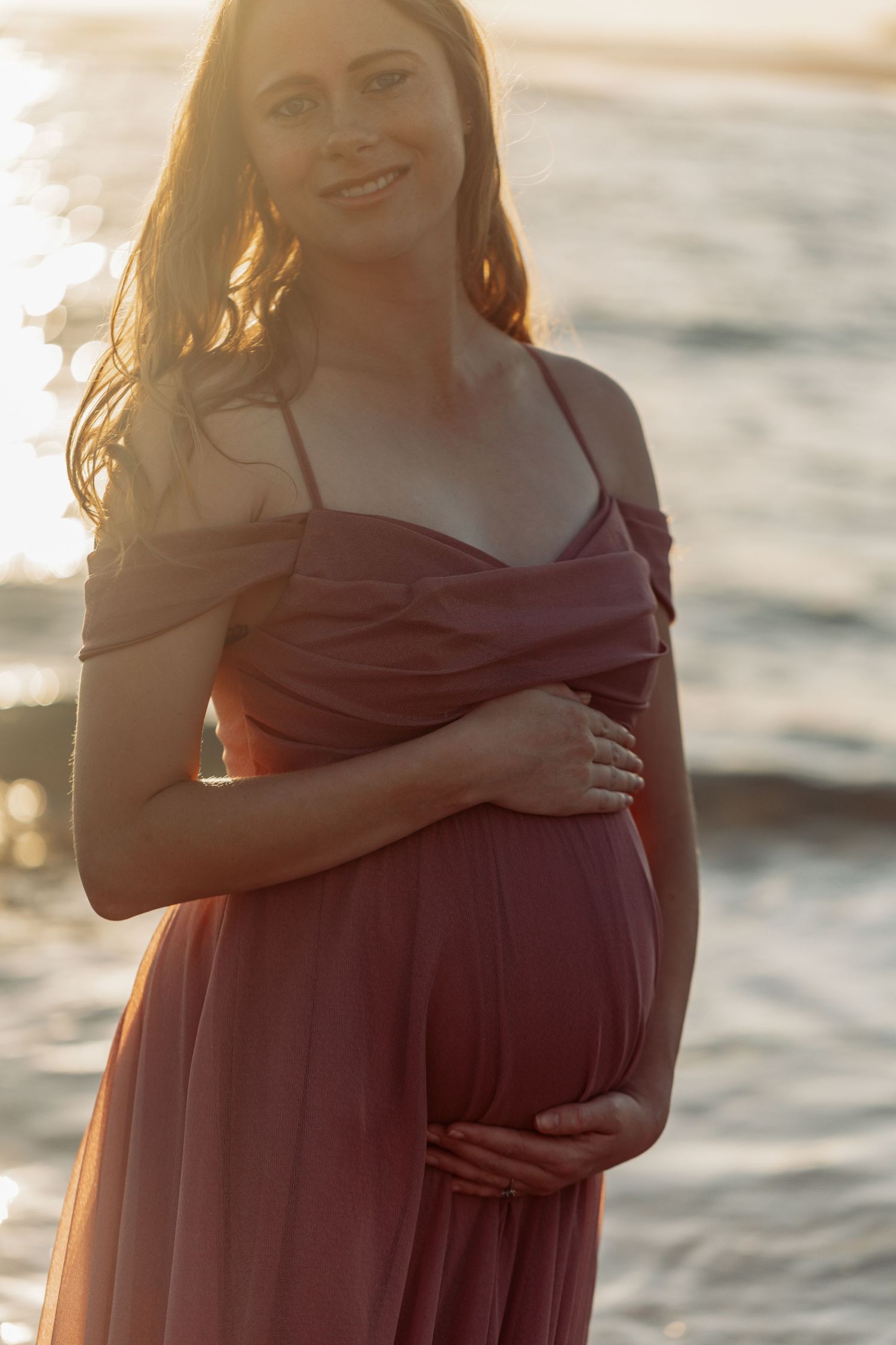 A pregnant woman in a burgundy dress stands by the ocean at sunset with golden light illuminating her silhouette.