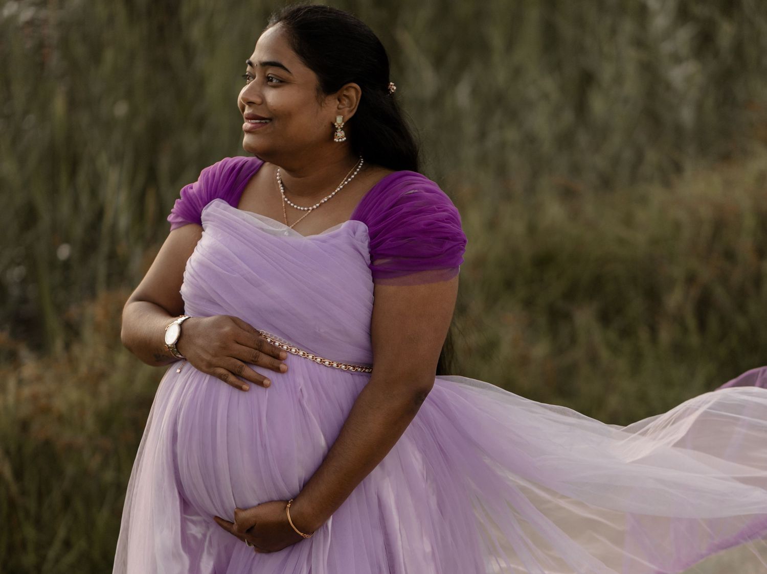 A woman in a flowing lavender gown cradles her baby bump while standing in a natural outdoor setting during sunset.