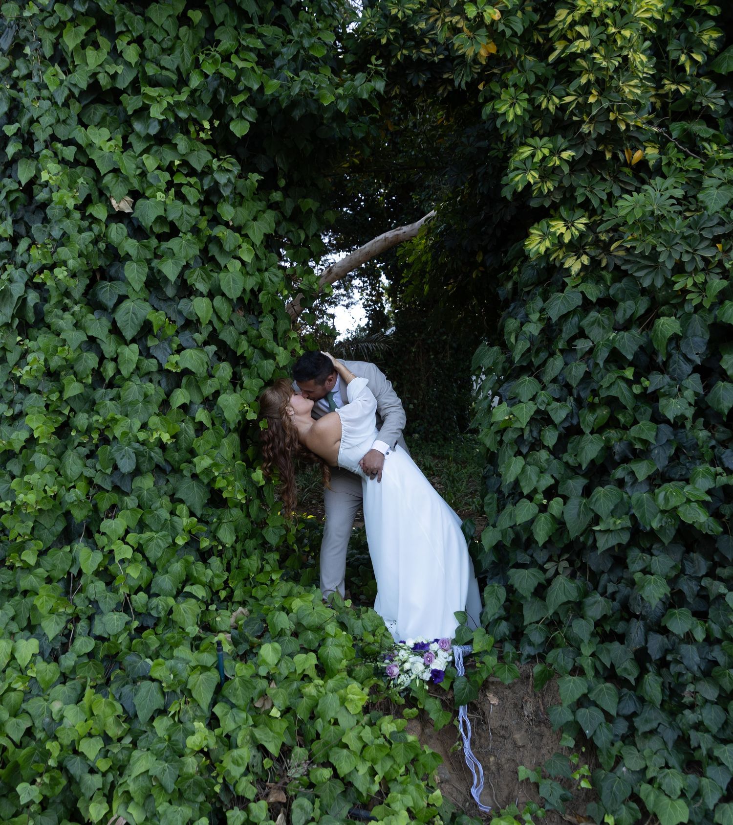 Bride and groom sharing a romantic kiss surrounded by lush green ivy walls in an enchanting outdoor wedding setting.